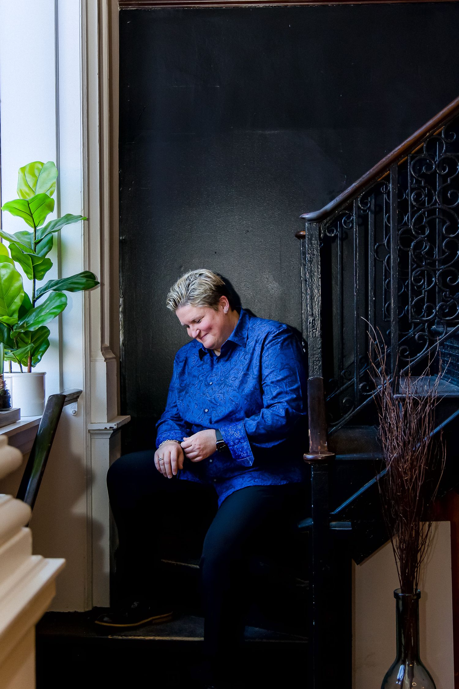Moody portrait of a person in blue sitting on dark stairs near a houseplant.