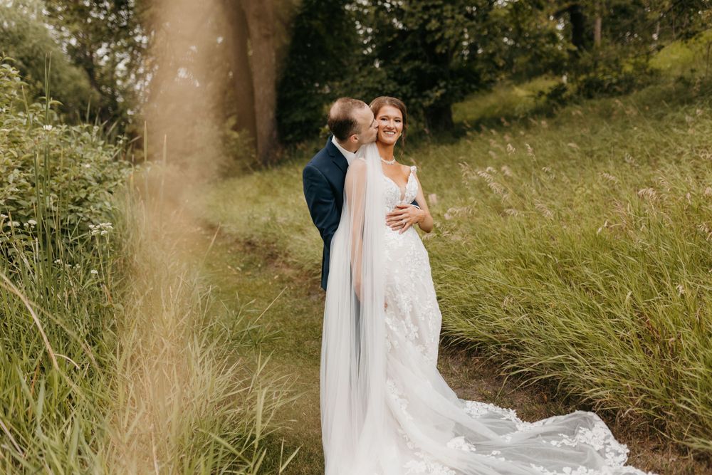 Bride in lace gown with cathedral veil standing with groom in nature setting with tall green grass.
