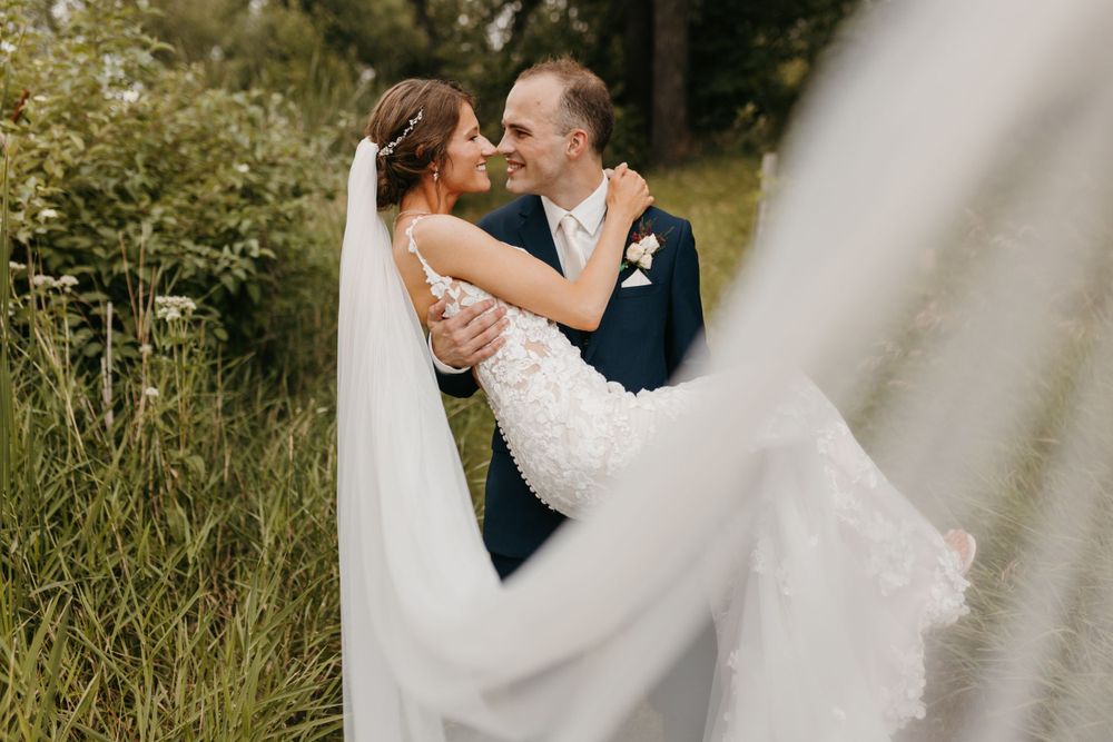 Groom lifting bride in lace wedding dress outdoors near tall grass, veil flowing in foreground.