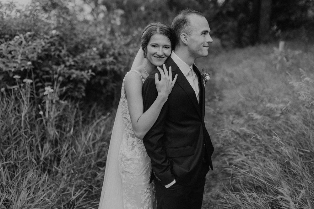 Black and white photo of bride leaning on groom's back smiling in outdoor nature setting.
