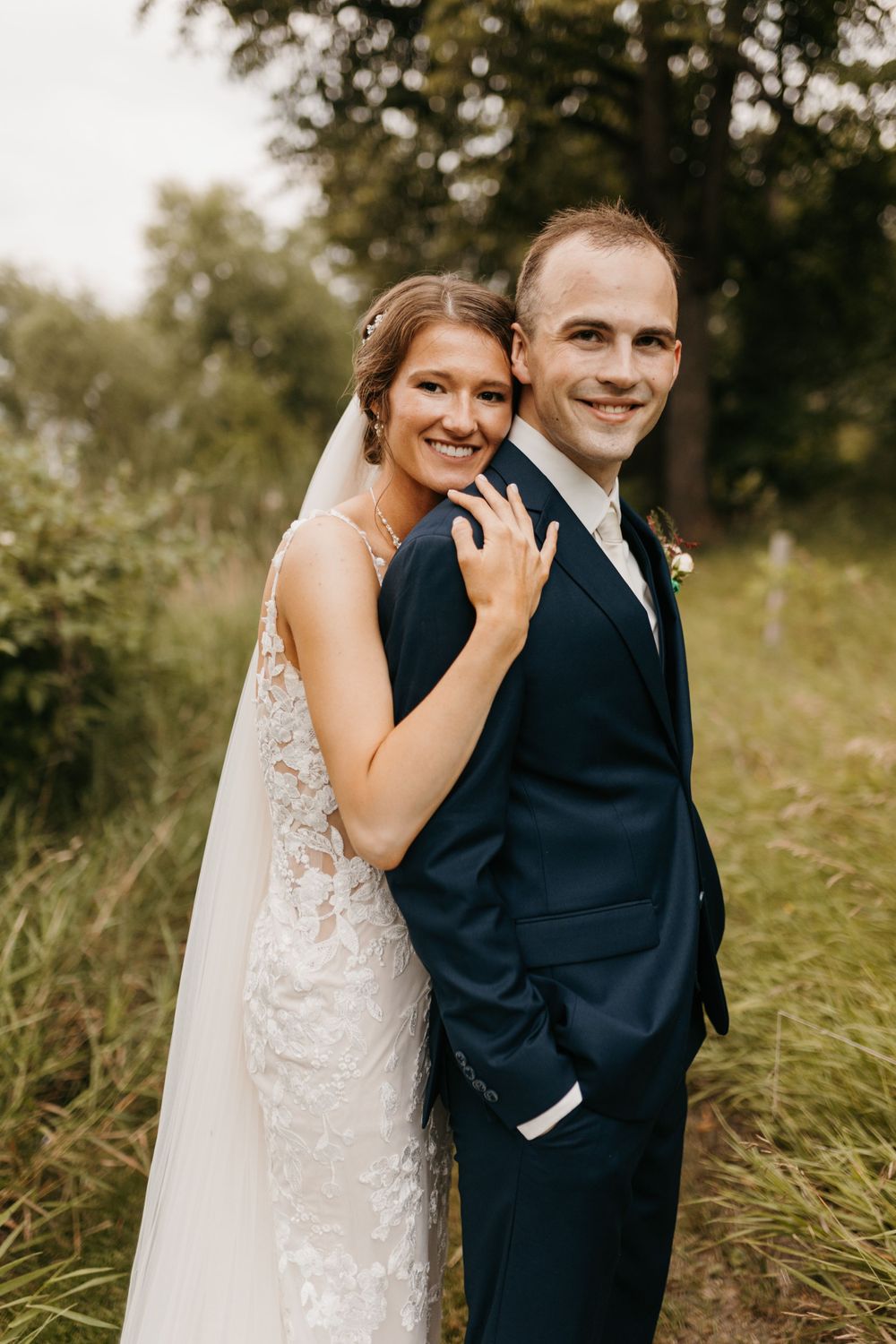 Bride in lace gown hugging groom in navy suit from behind, both smiling outdoors on wedding day.