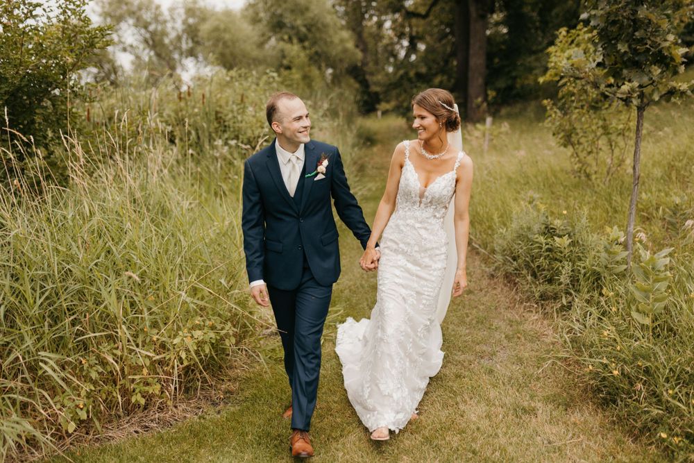 Bride and groom walk hand in hand through a lush green meadow path on their wedding day.