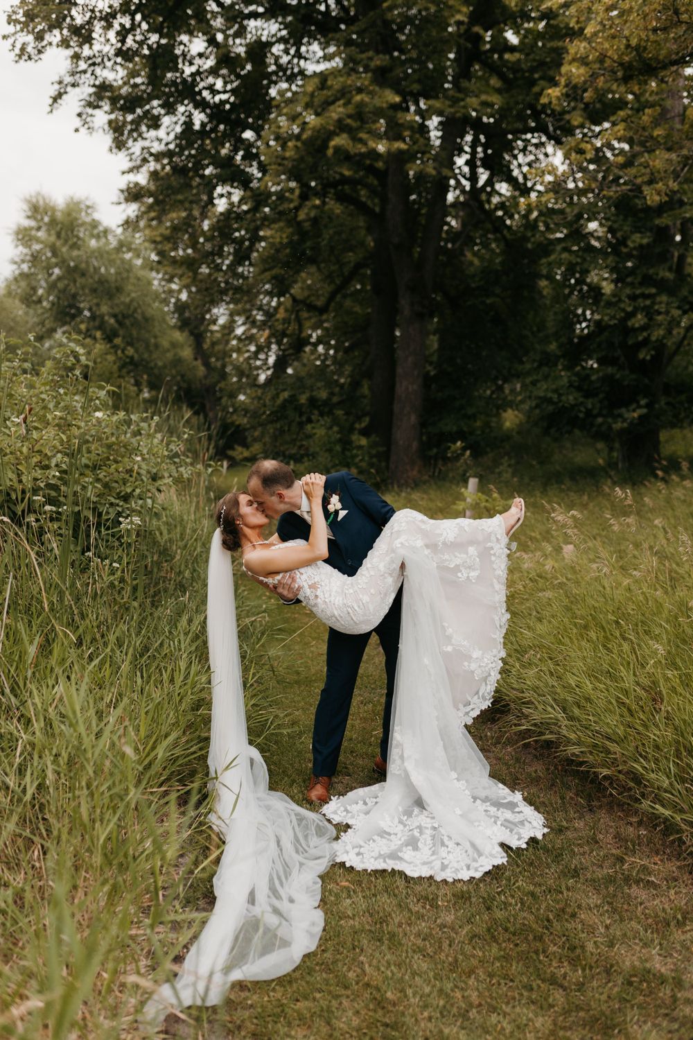 Groom dipping bride for a romantic kiss outdoors in green field, bride's veil trailing on ground.