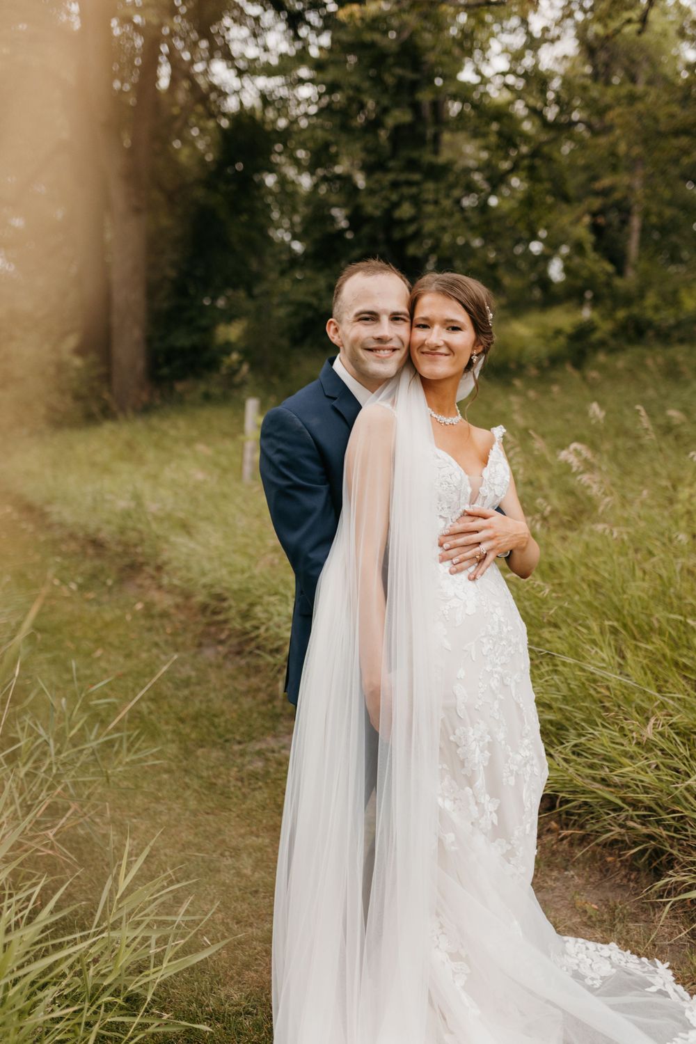 Happy couple posing together outdoors, groom in navy suit hugging bride in lace gown with flowing veil.