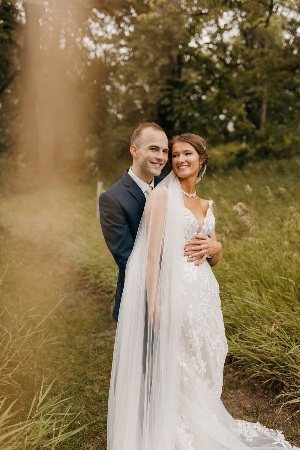 Bride and groom embracing outdoors in golden sunlight, bride wearing lace wedding dress with long veil.