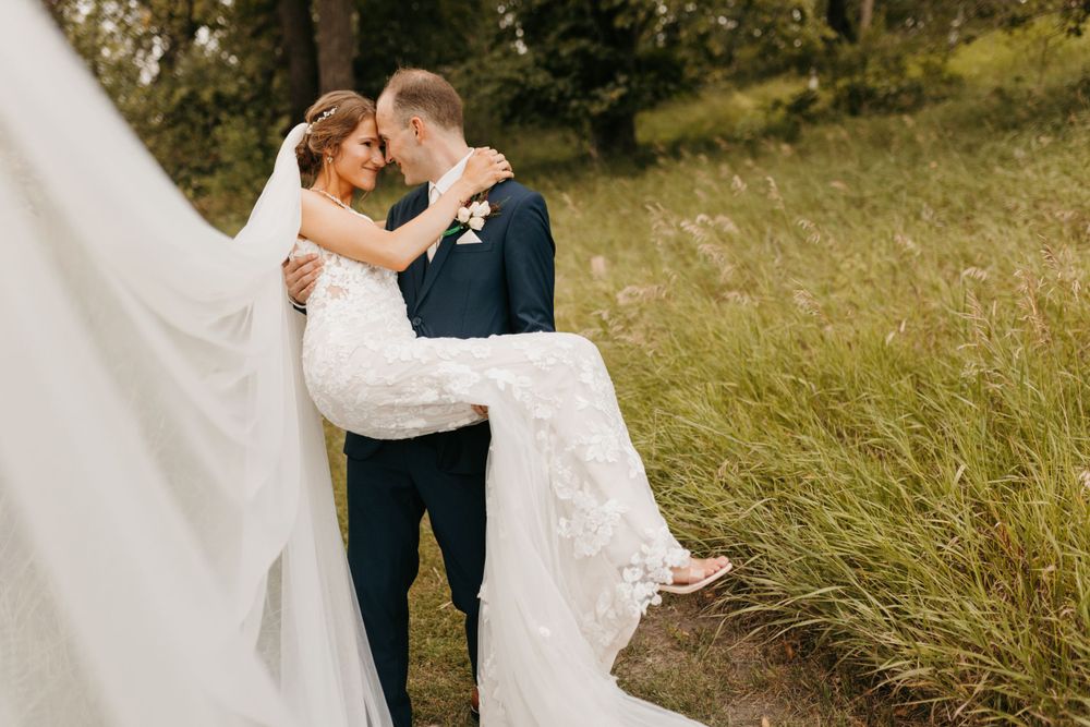 Groom lifting smiling bride in lace wedding dress outdoors beside tall grass on wedding day.