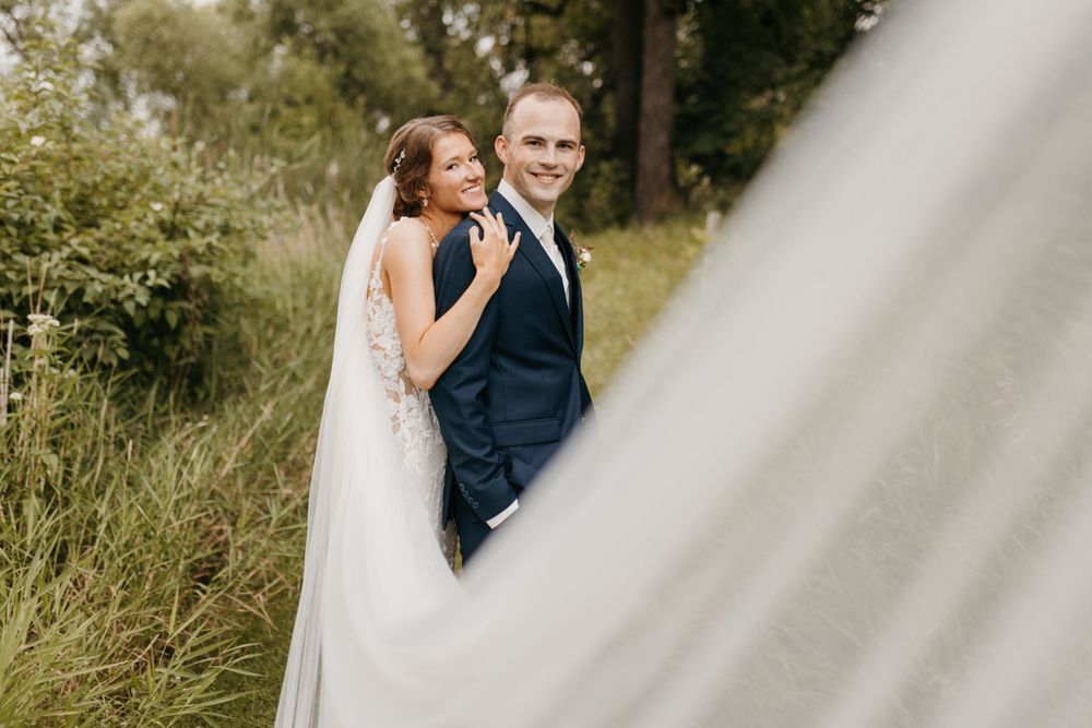 Bride and groom posing together outdoors with flowing veil in foreground and lush greenery behind.