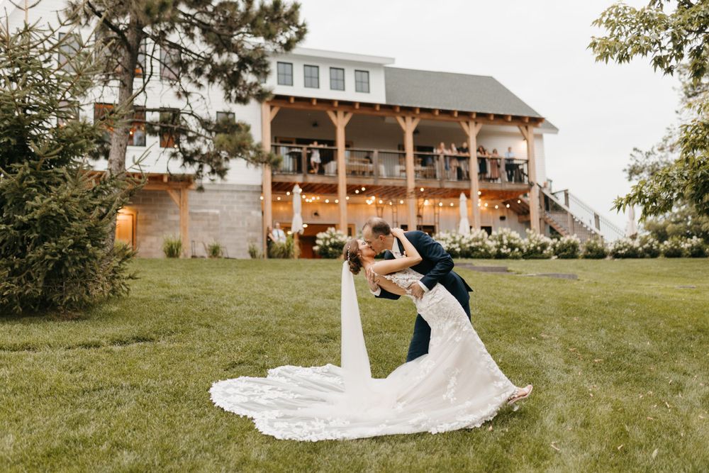 Groom dips bride in white gown on green lawn in front of elegant wedding venue with string lights.