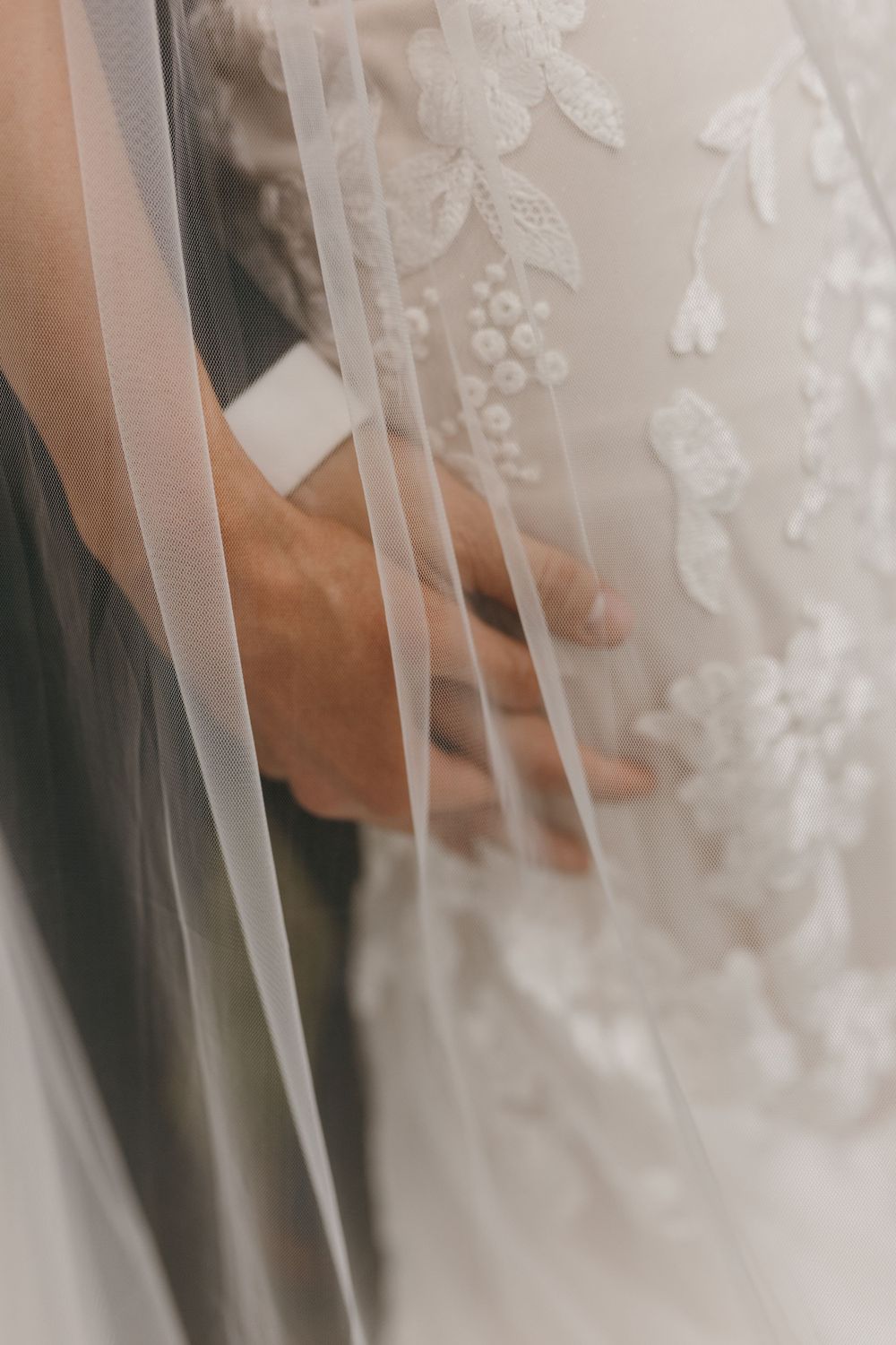 Close-up of groom's hand resting on bride's lace wedding gown with flowing veil.
