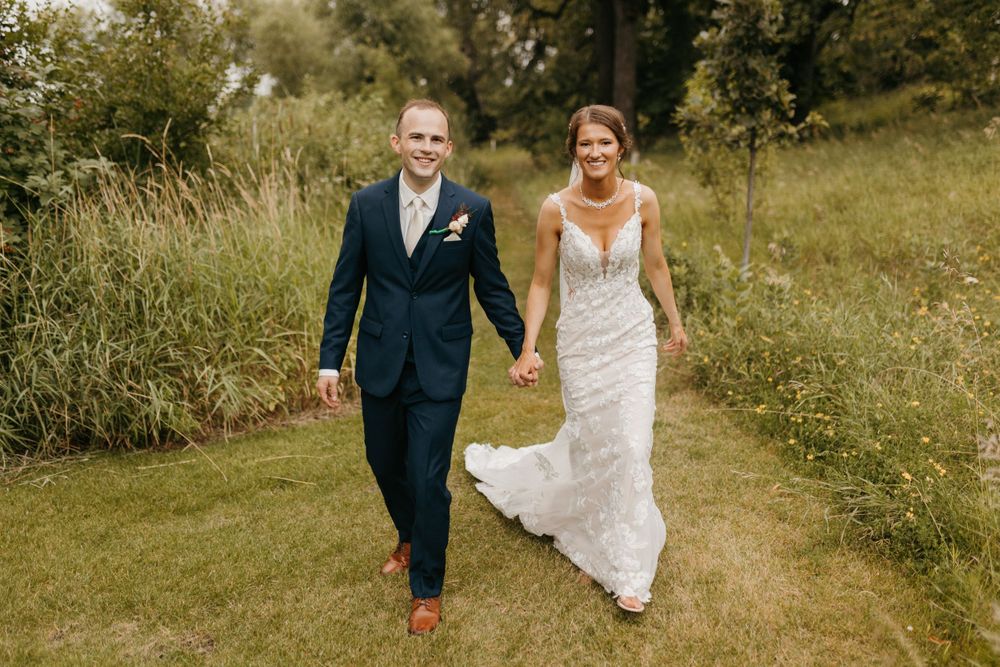 Happy newlyweds stroll along a scenic green path, bride in lace gown and groom in navy suit.