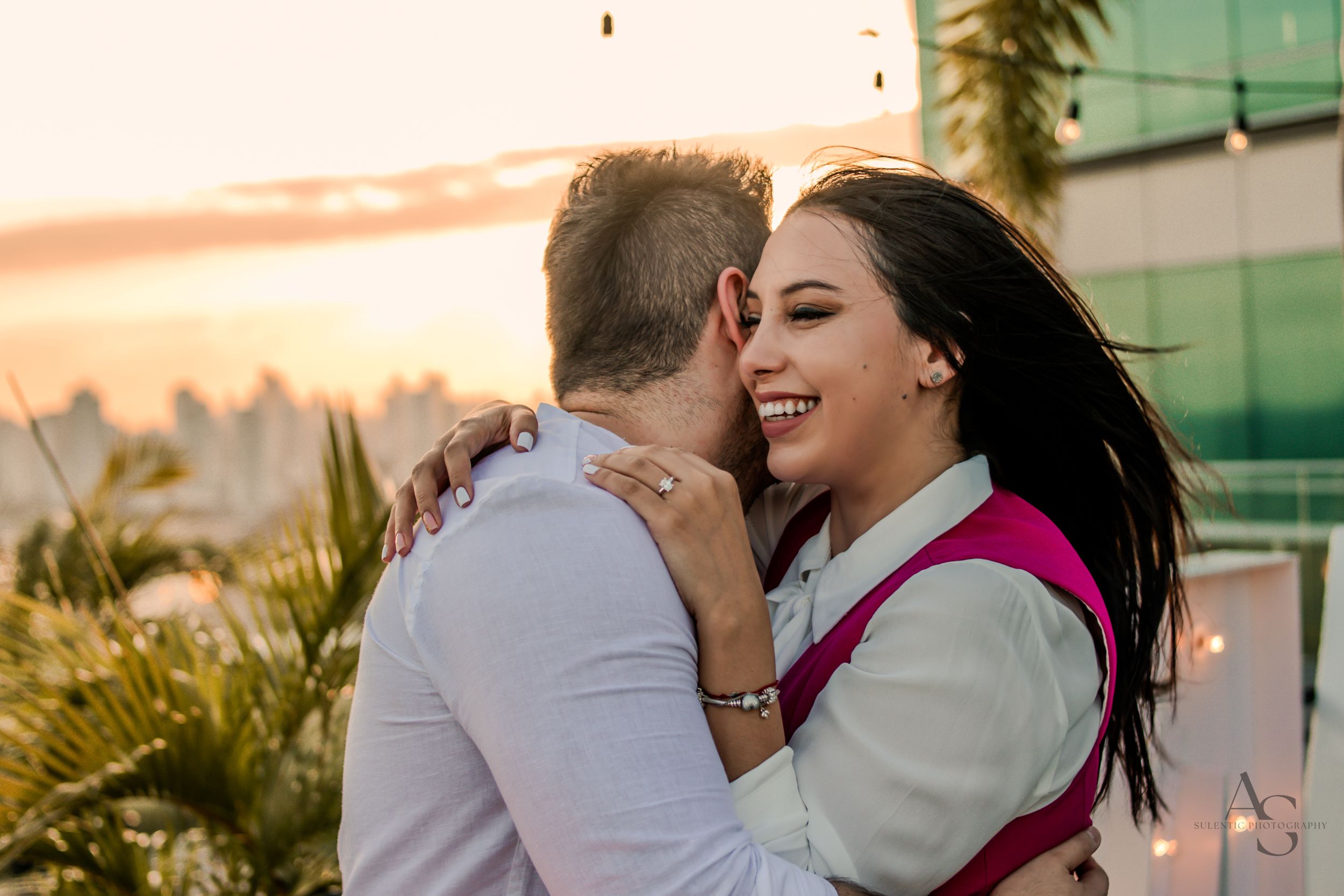 A couple embraces and laughs together on a rooftop during golden hour with palm trees visible.