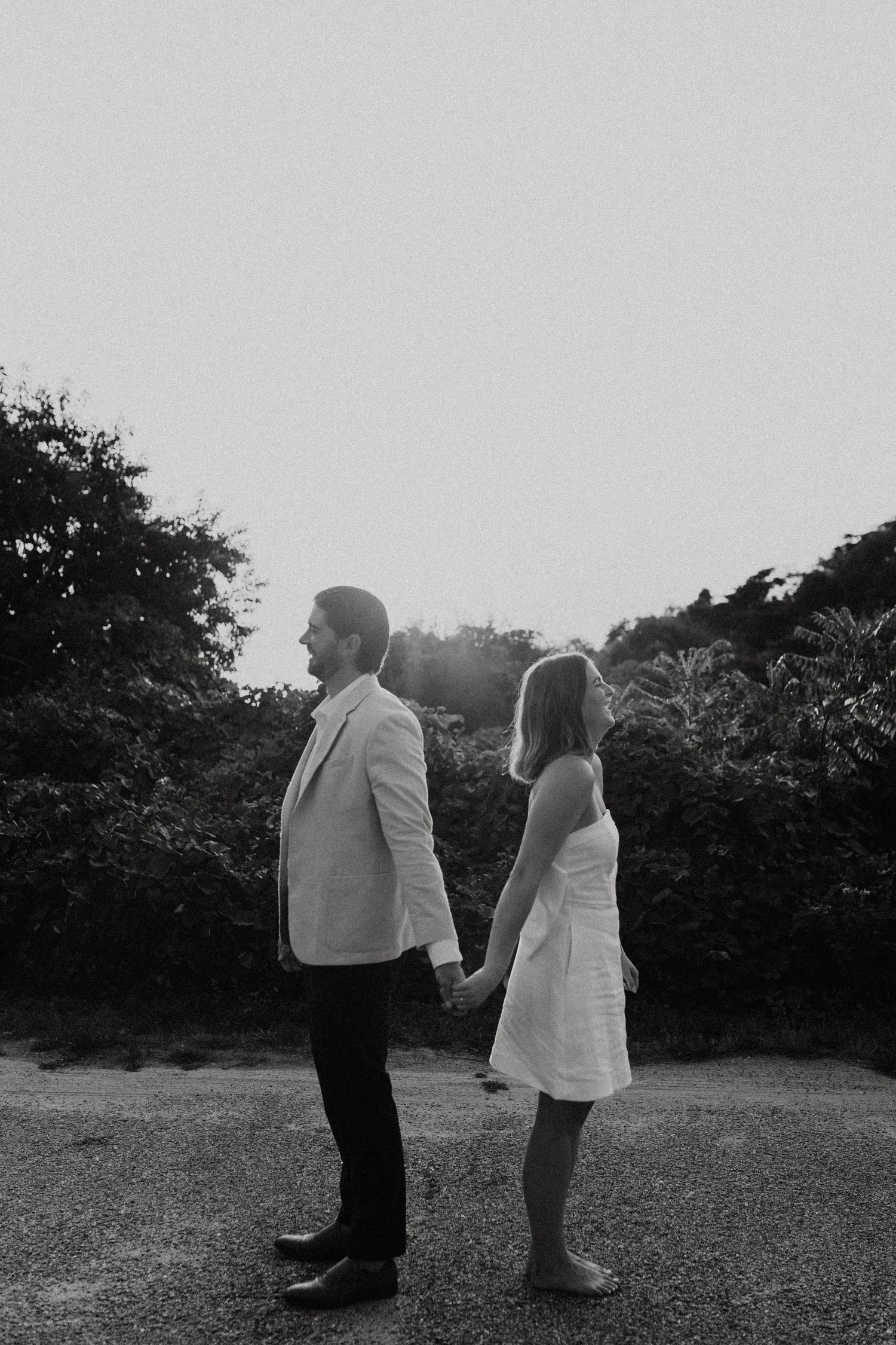 Couple holding hands back-to-back on gravel path surrounded by lush greenery in black and white.
