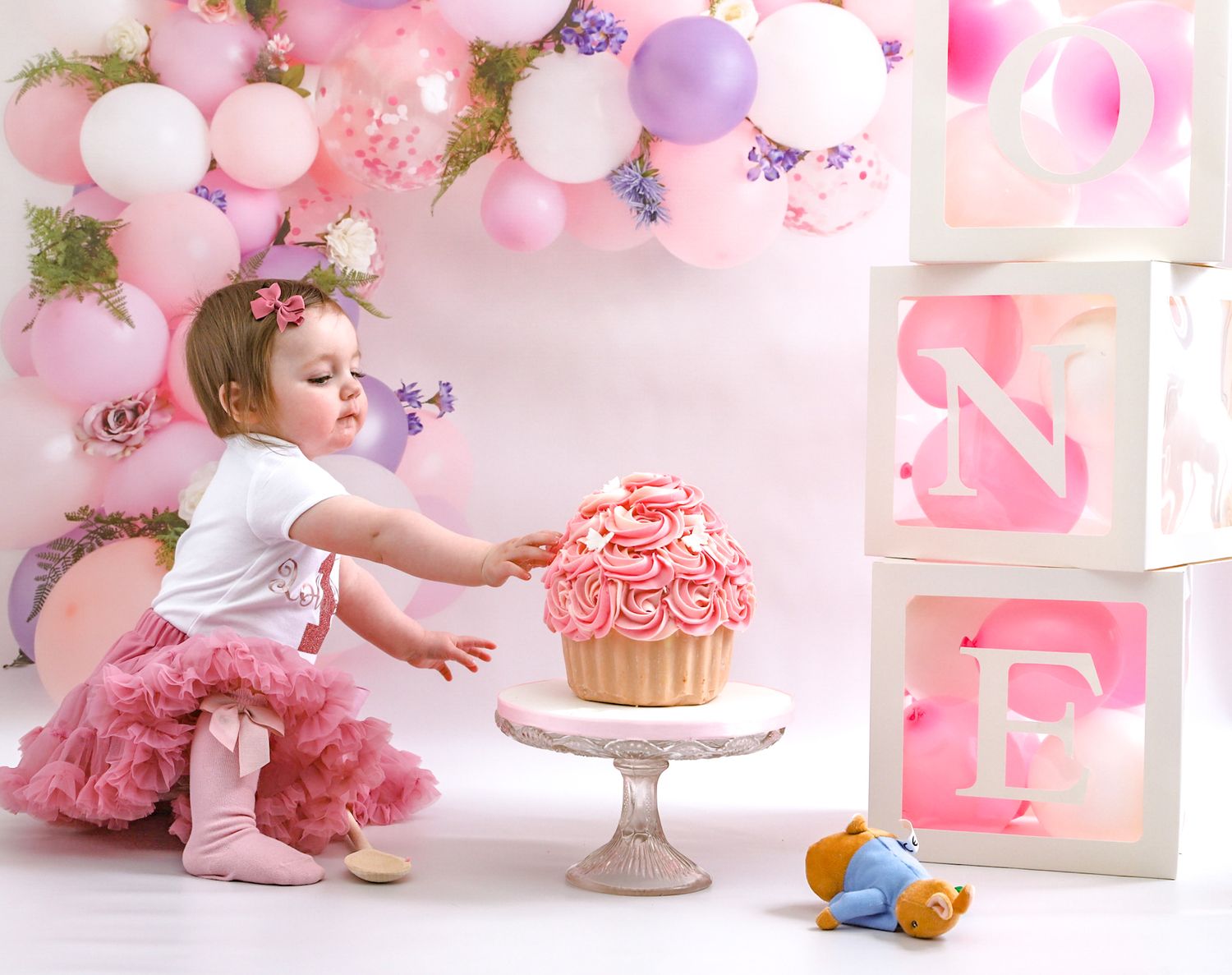 Pink and purple balloon arch decorates a cake smash photo session with a large cupcake on a pedestal.
