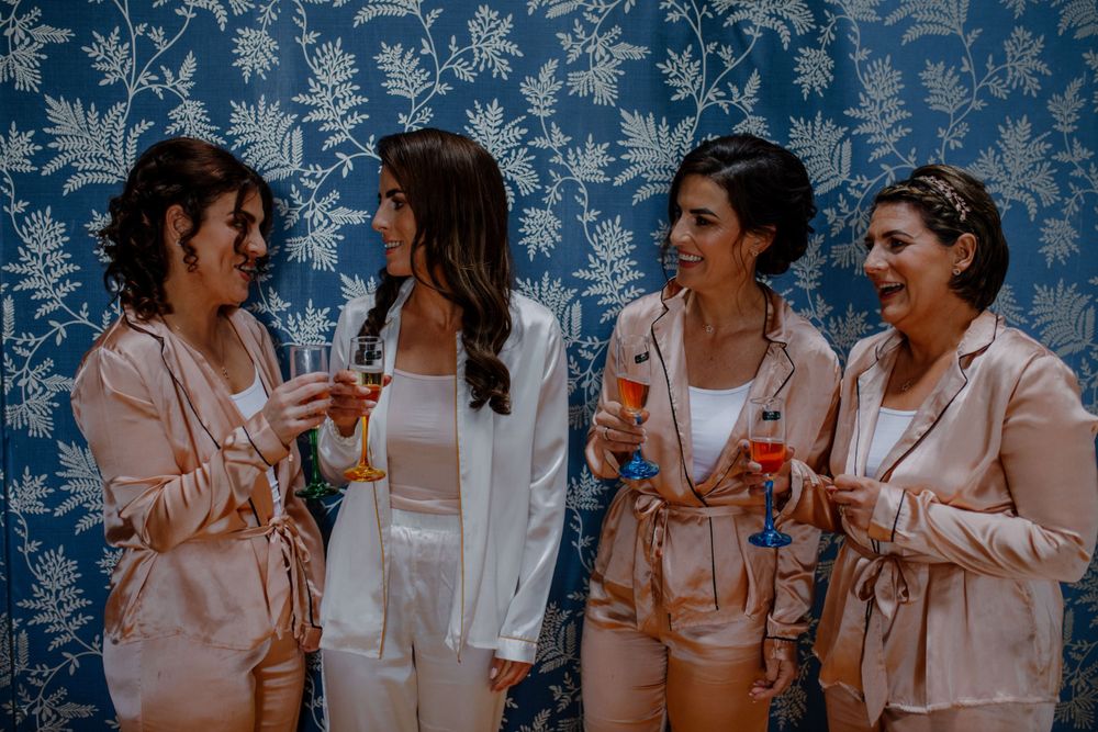 Four women in matching satin pajama sets cheer with colorful drinks against a floral-patterned backdrop, exuding a festive atmosphere.