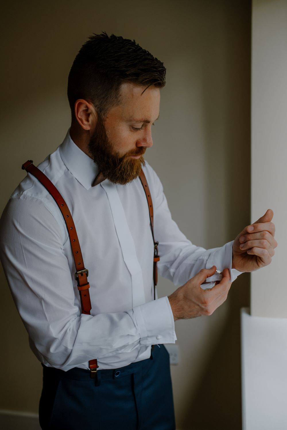 A man in a white dress shirt adjusts his cufflinks, wearing brown leather suspenders over dark pants, in a softly lit room.