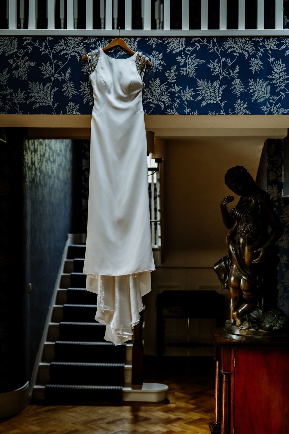 A sleek white wedding dress hangs gracefully against a backdrop of blue floral wallpaper, above a dark wooden statue and staircase.