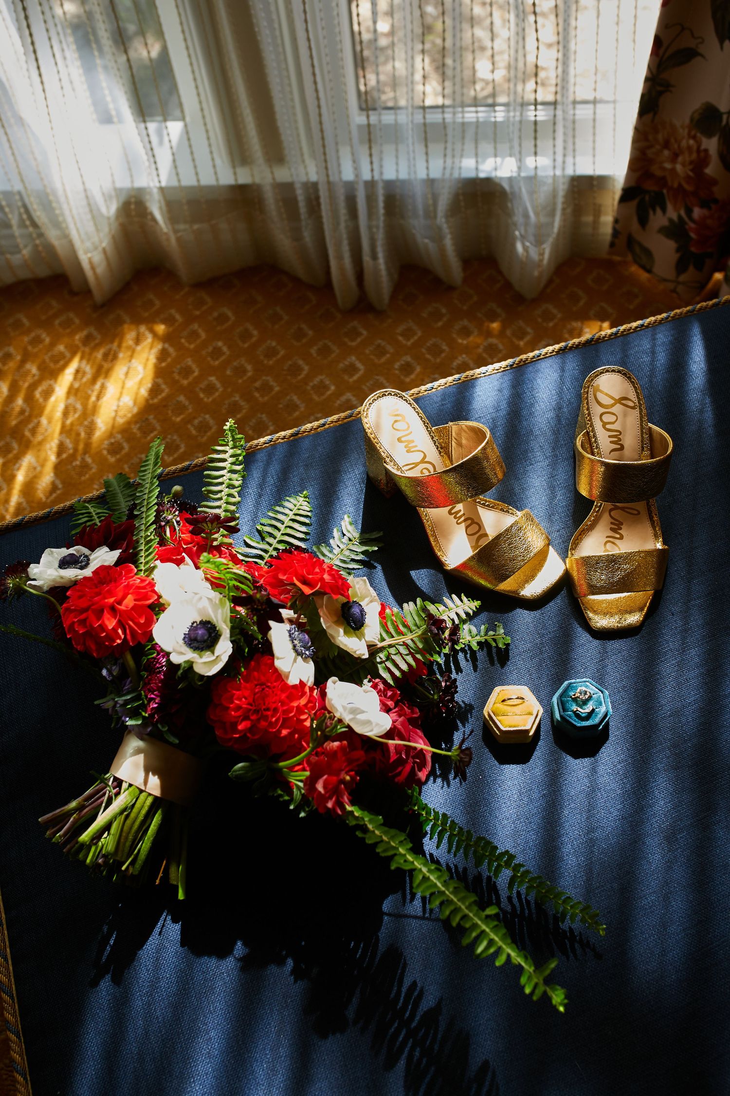 Wedding details including red and white bouquet with gold shoes on blue fabric in sunlight.