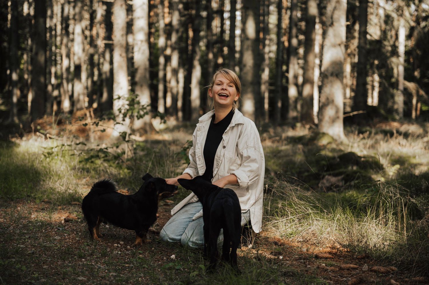 A smiling woman in a white jacket plays with a black dog in a sunlit forest clearing.