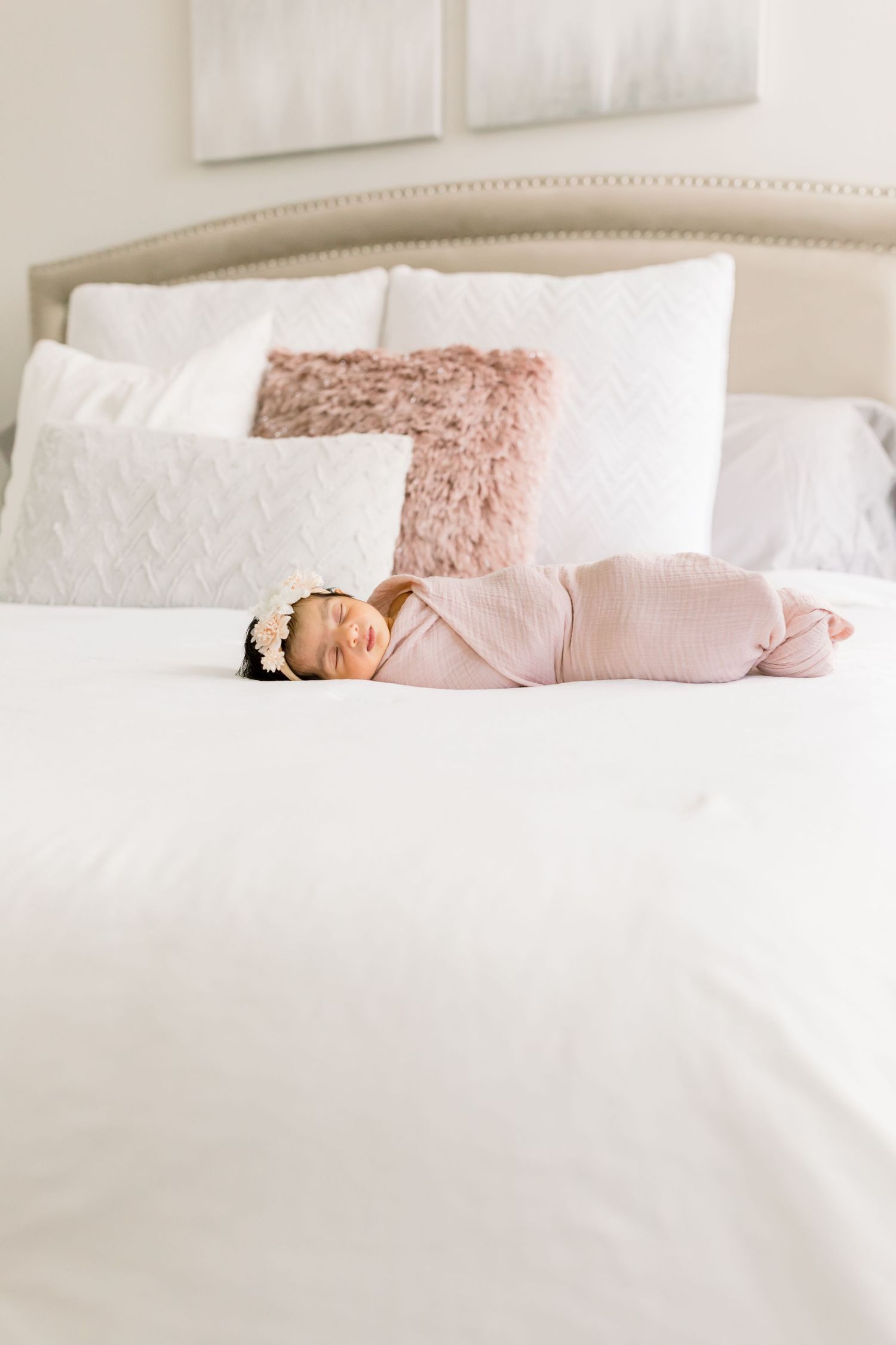 Sleeping baby in pink swaddle on white bedding with plush pillows.