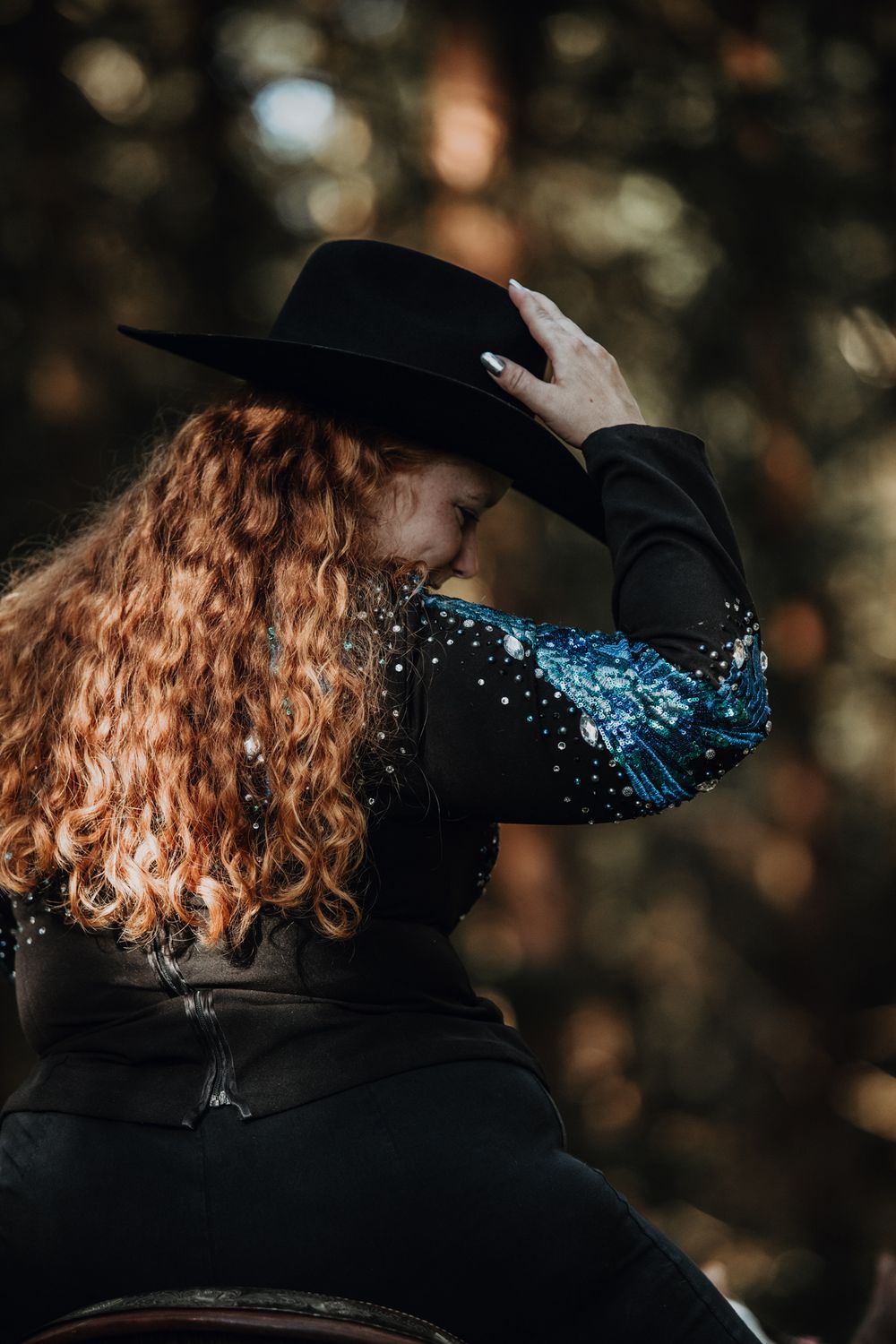Equestrian wearing black hat and sparkly jacket with flowing red hair against forest backdrop.