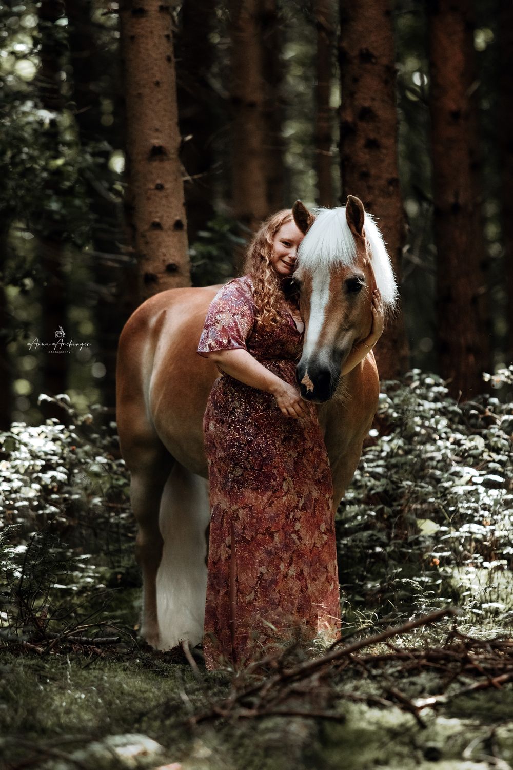 Beautiful haflinger horse stands next to rider in floral dress in mystical forest setting.