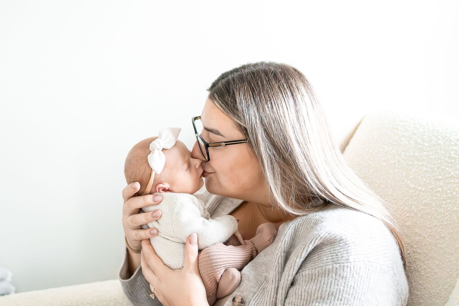 A caregiver in glasses and gray sweater gently kisses their sleeping newborn wearing a white bow.