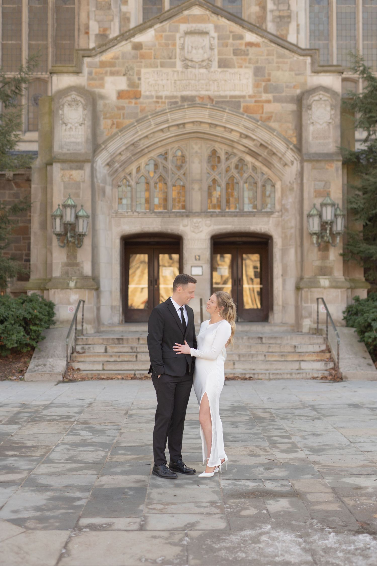 A bride and groom standing together in front of a large, stone building with arched doorways. Ann Arbor Law Quad Engagement Photos