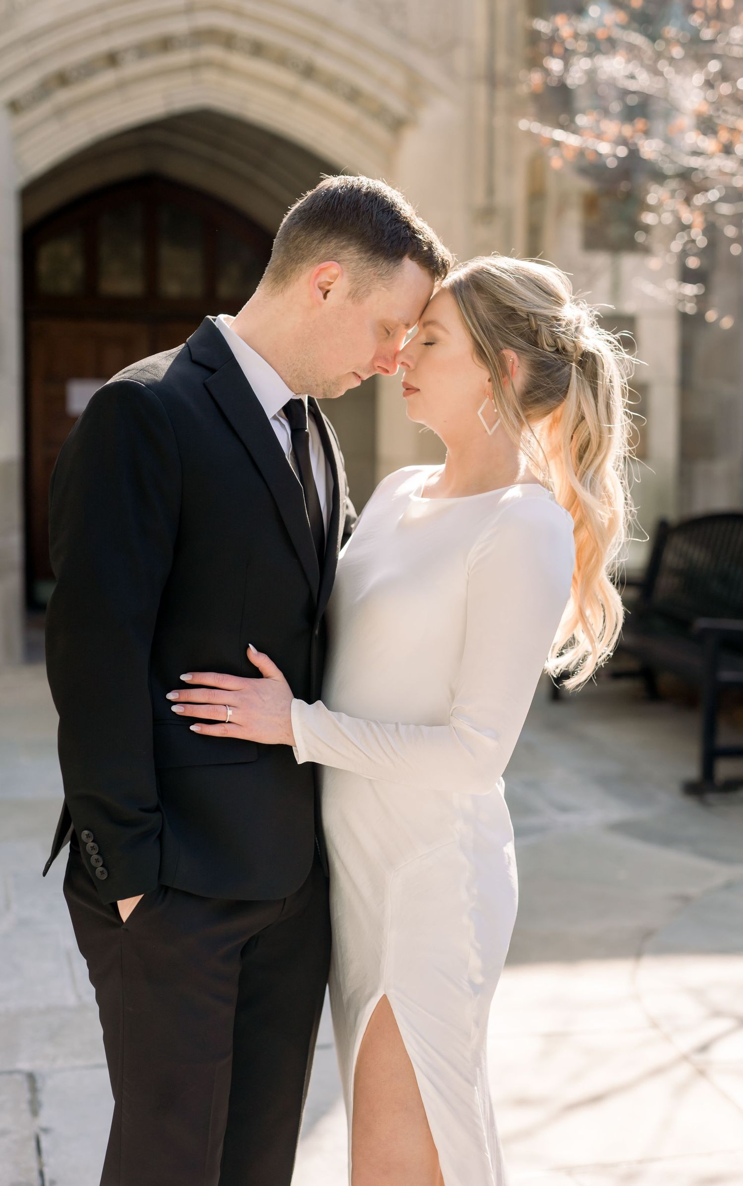 A bride and groom embracing each other in front of a large, stone building with arched doorways. Ann Arbor Law Quad Engagement Photos