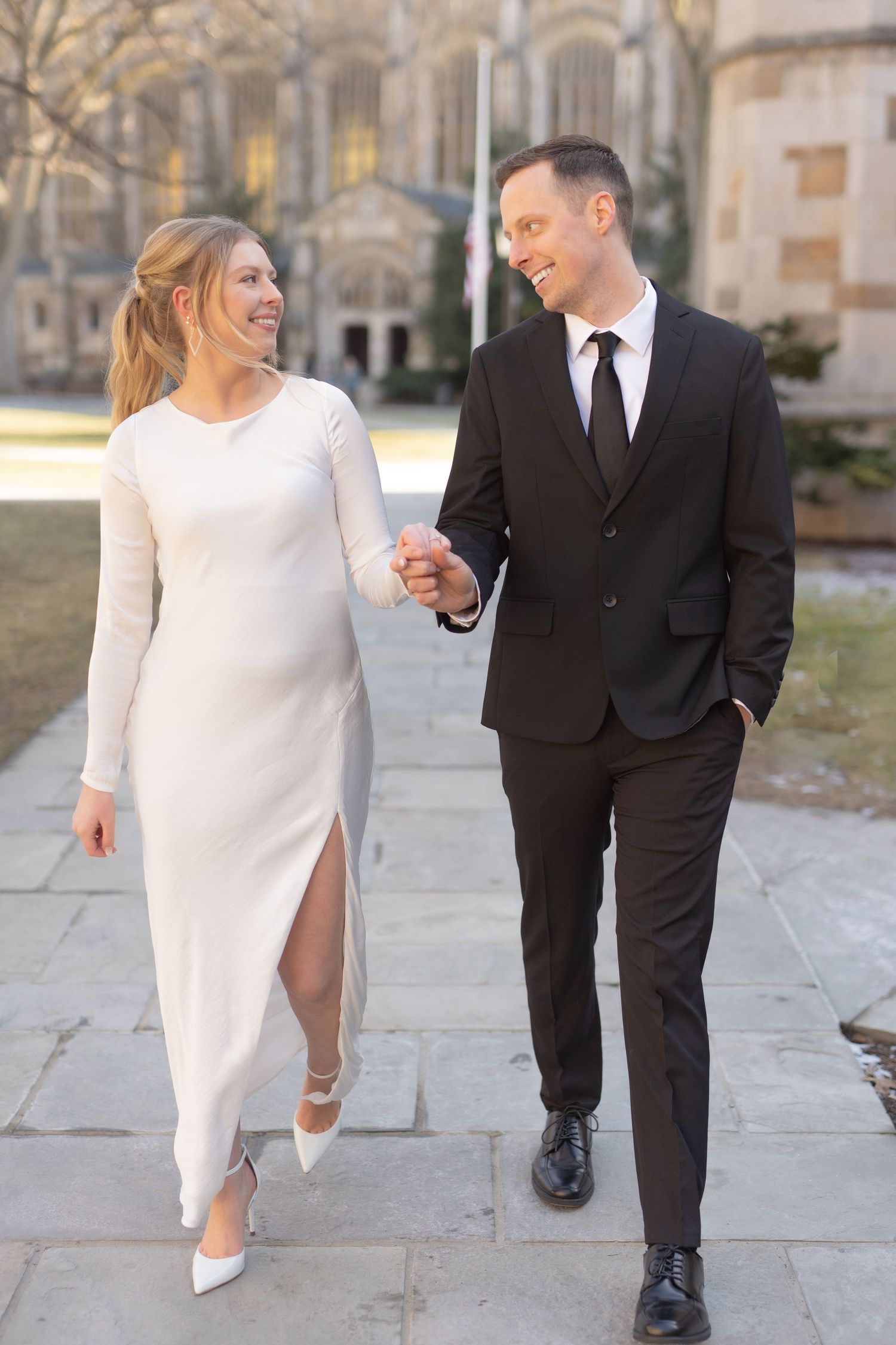 A couple walking together on a paved path in front of a large, stone building. Ann Arbor Law Quad Engagement Photos
