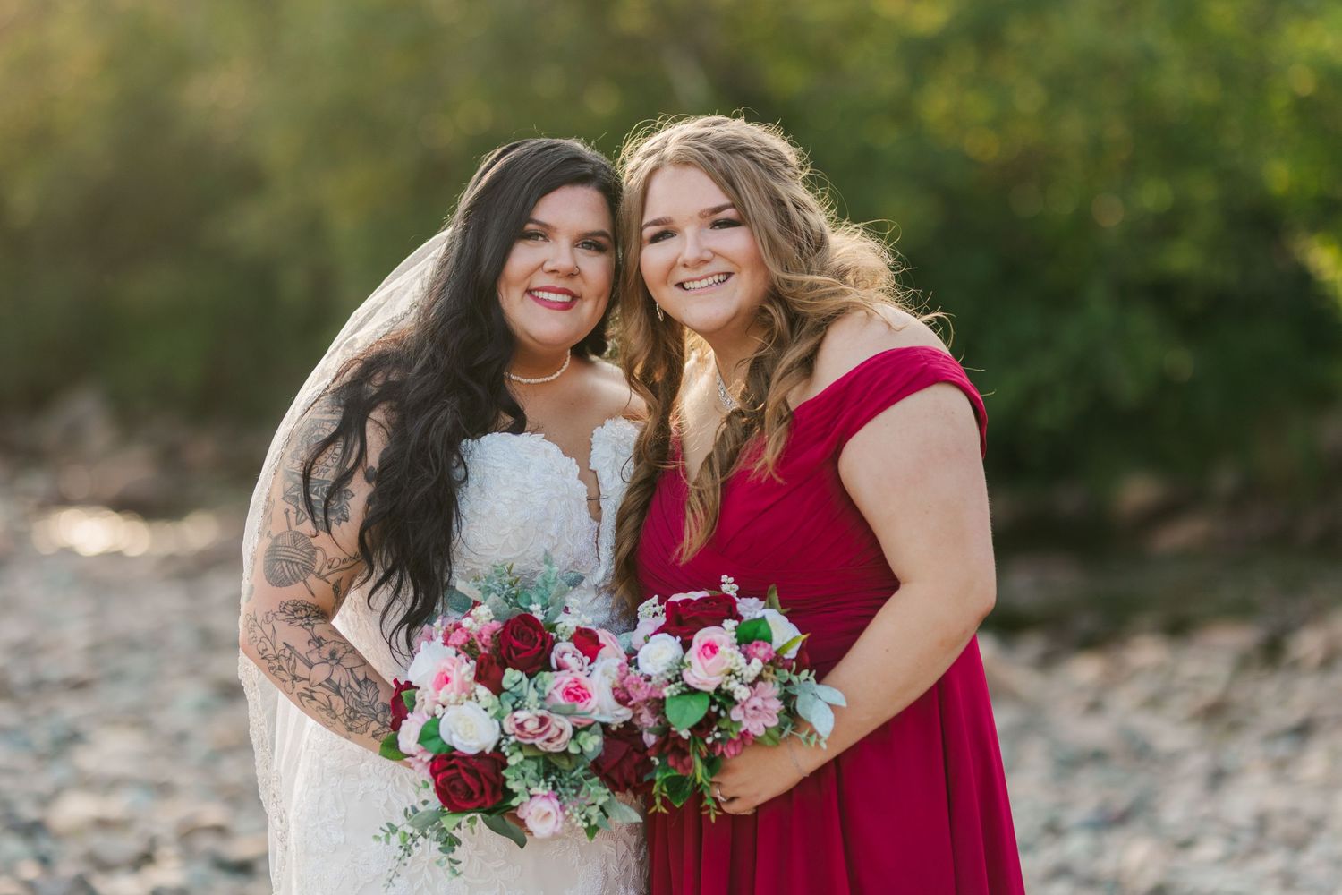 A bride in white and attendant in red hold pink and white bouquets while posing outdoors.