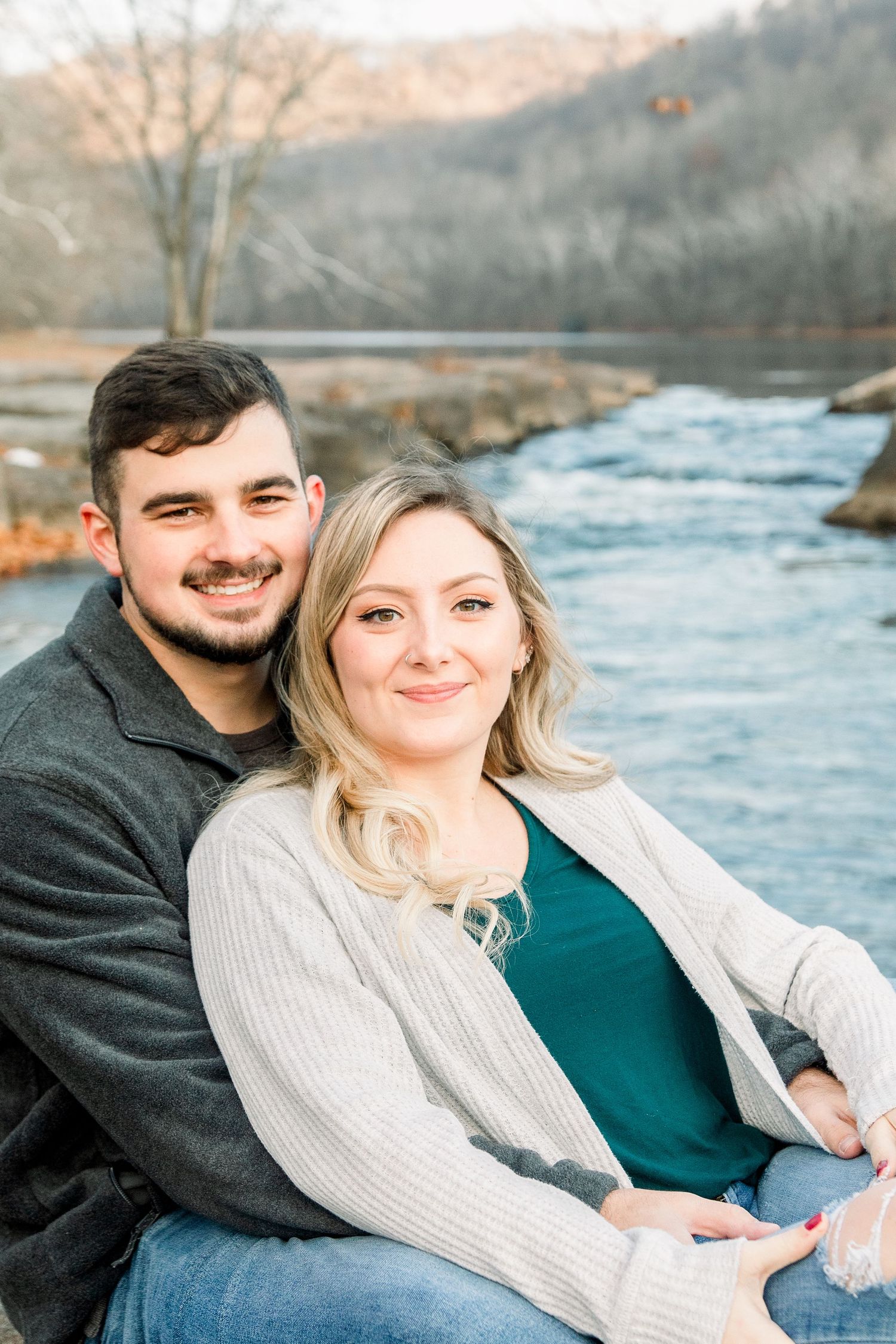 Couple sits by river during engagement photo session in casual winter attire.