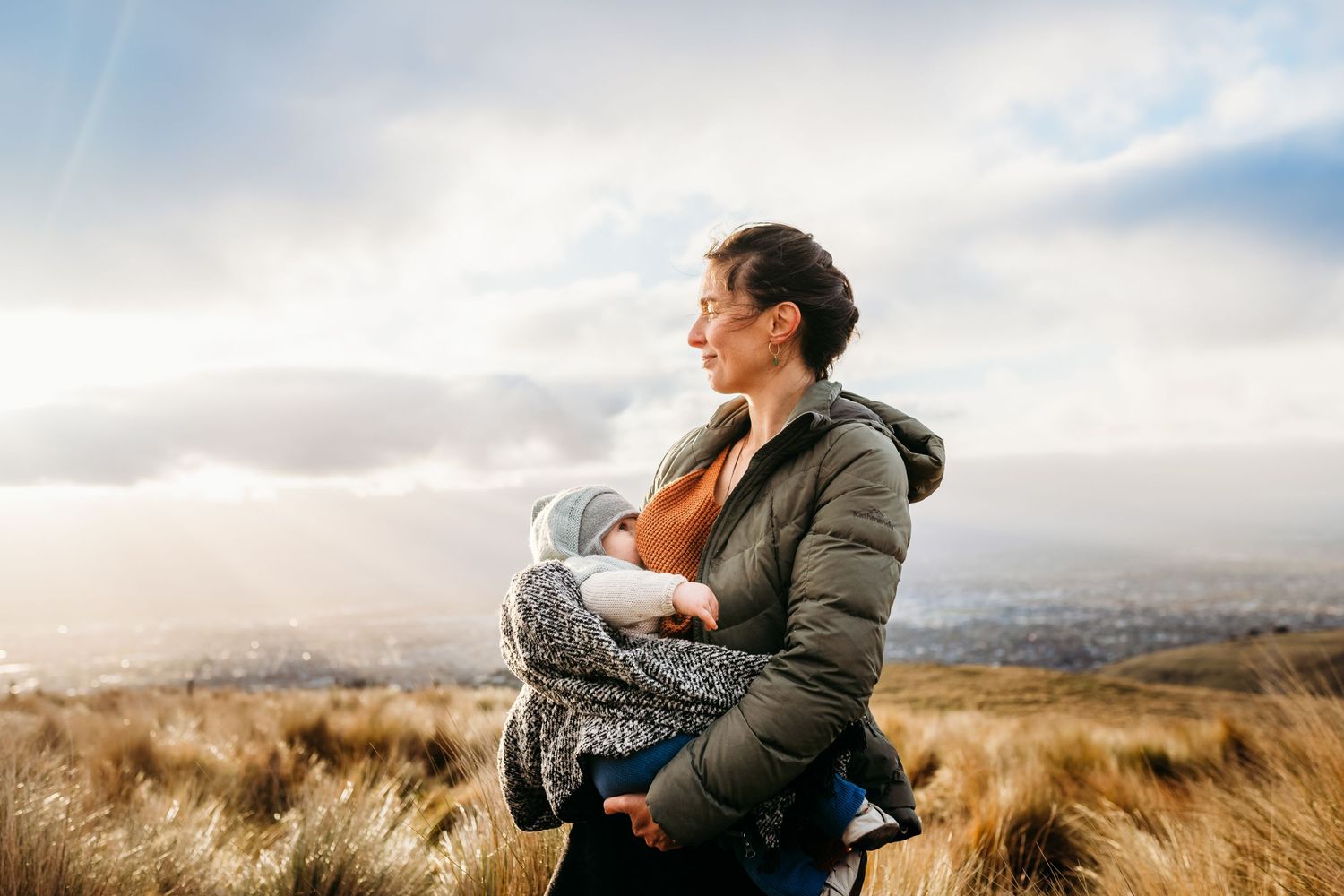 A person wearing a puffy jacket holds a baby while overlooking a scenic vista.
