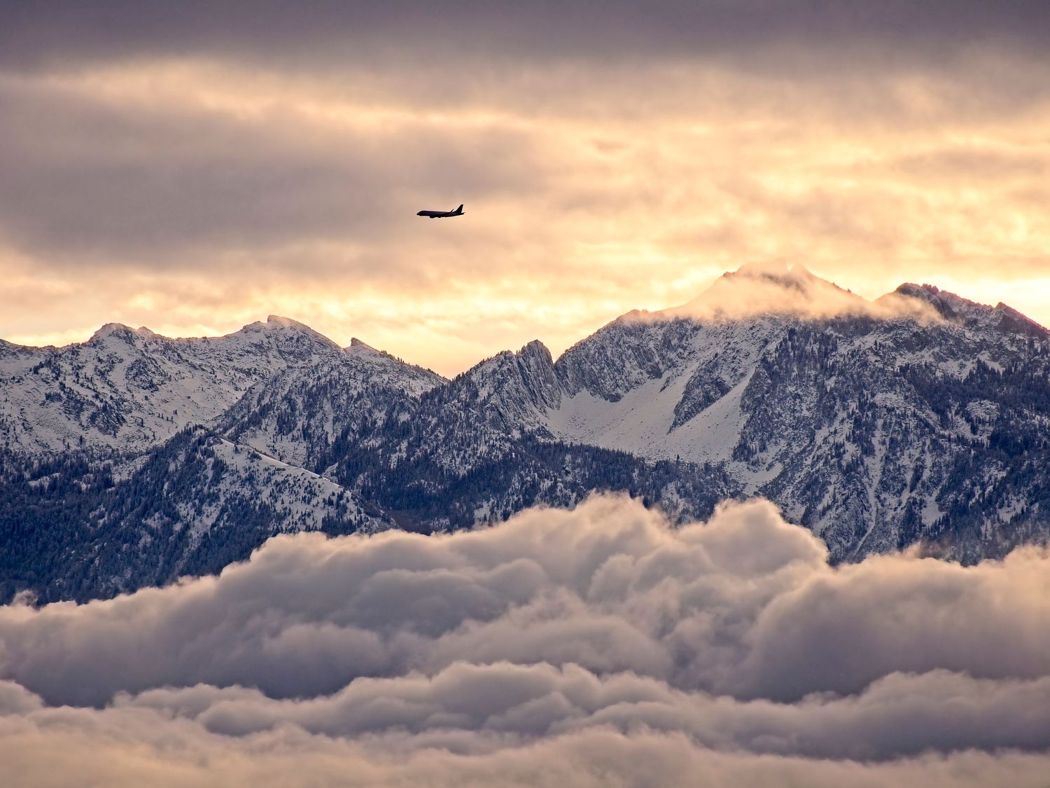 Mountains in the sunrise stand above the low rolling clouds while the clouds above catch the sunlight. A passenger airline flies above