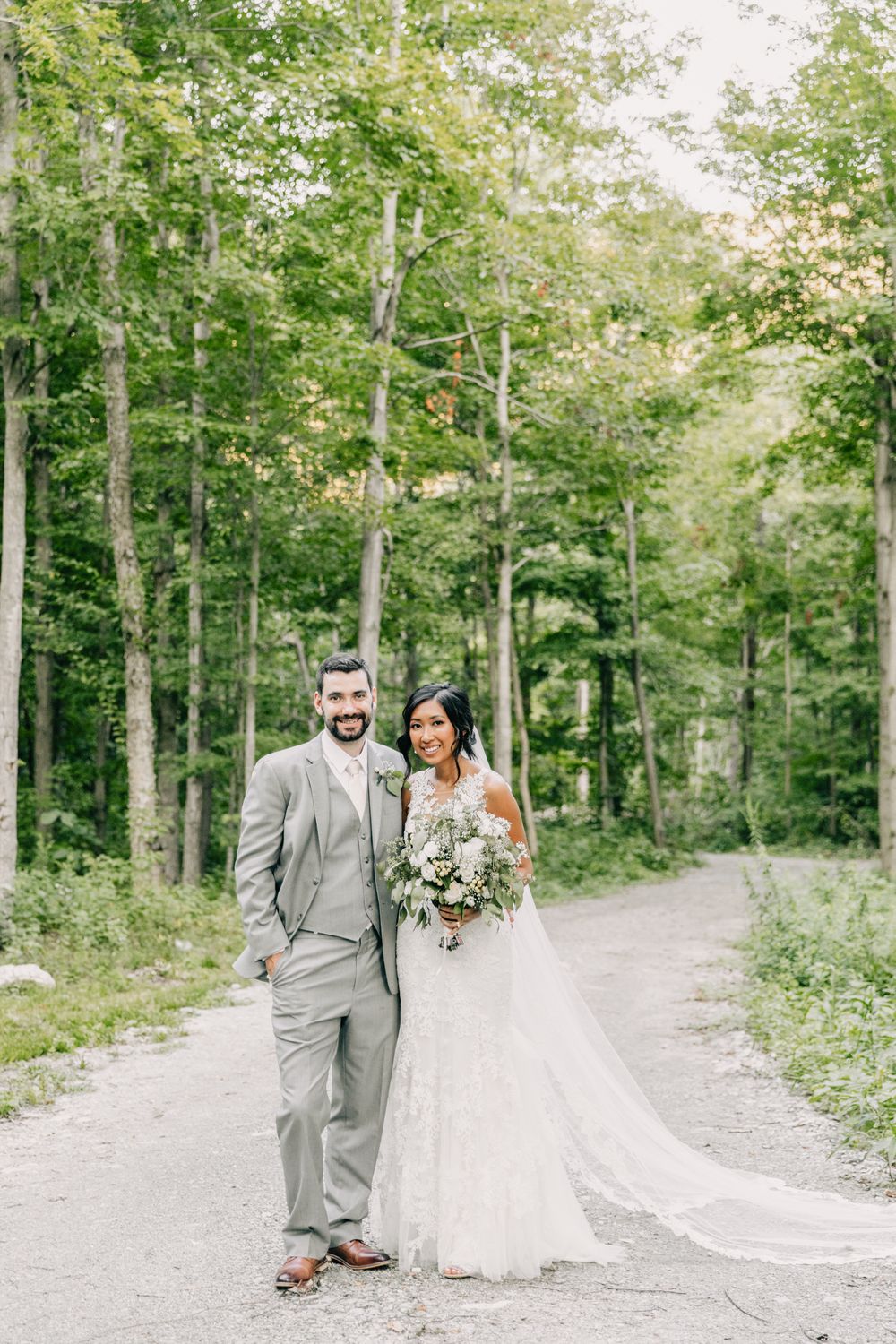 A newlywed couple poses on a wooded path, with the groom in grey suit and bride in lace gown.