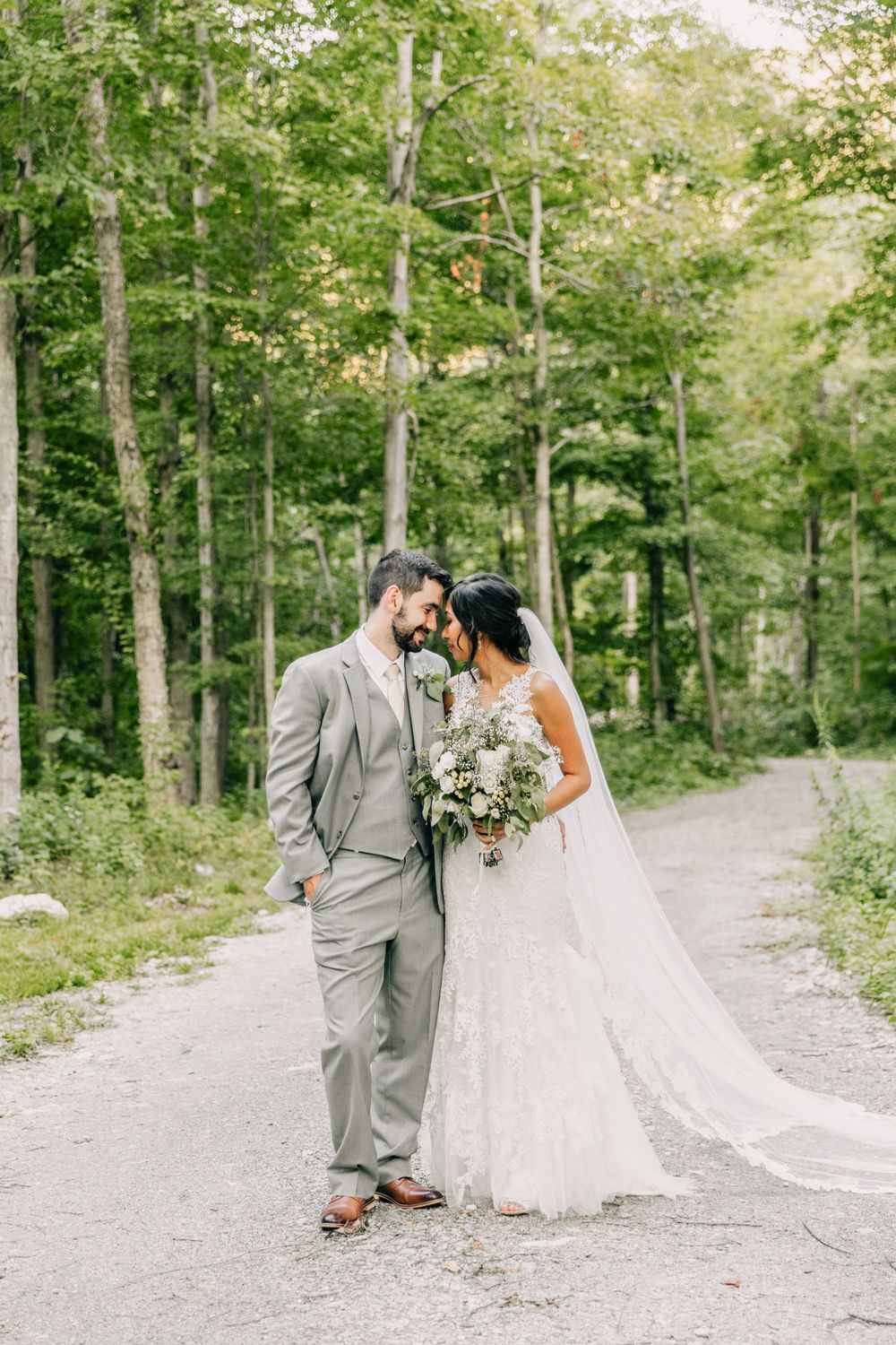 A romantic wedding portrait of a couple on a forest path, with the bride holding a delicate white bouquet.