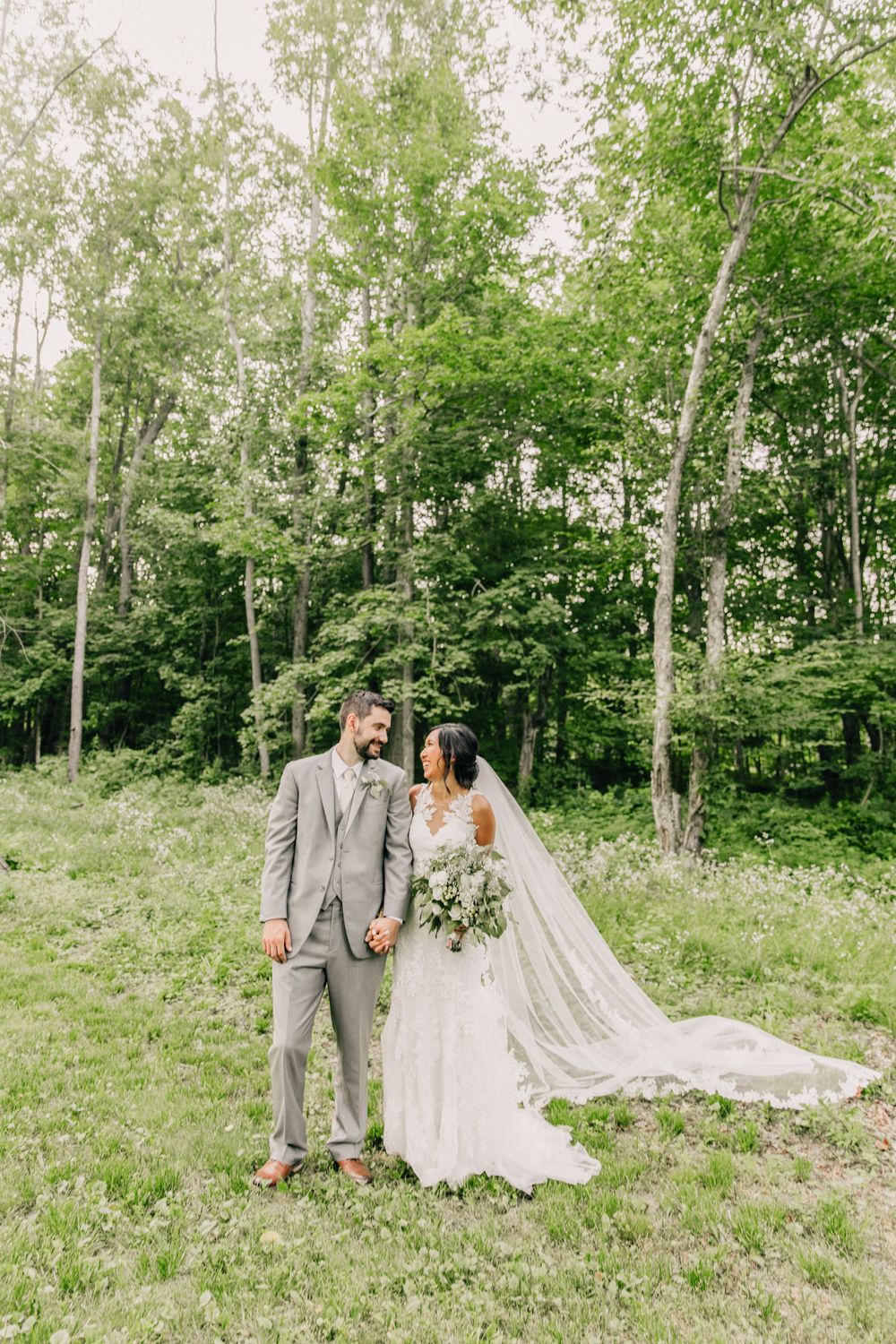 Wedding couple stands in a sunlit forest clearing with flowing veil and grey suit.