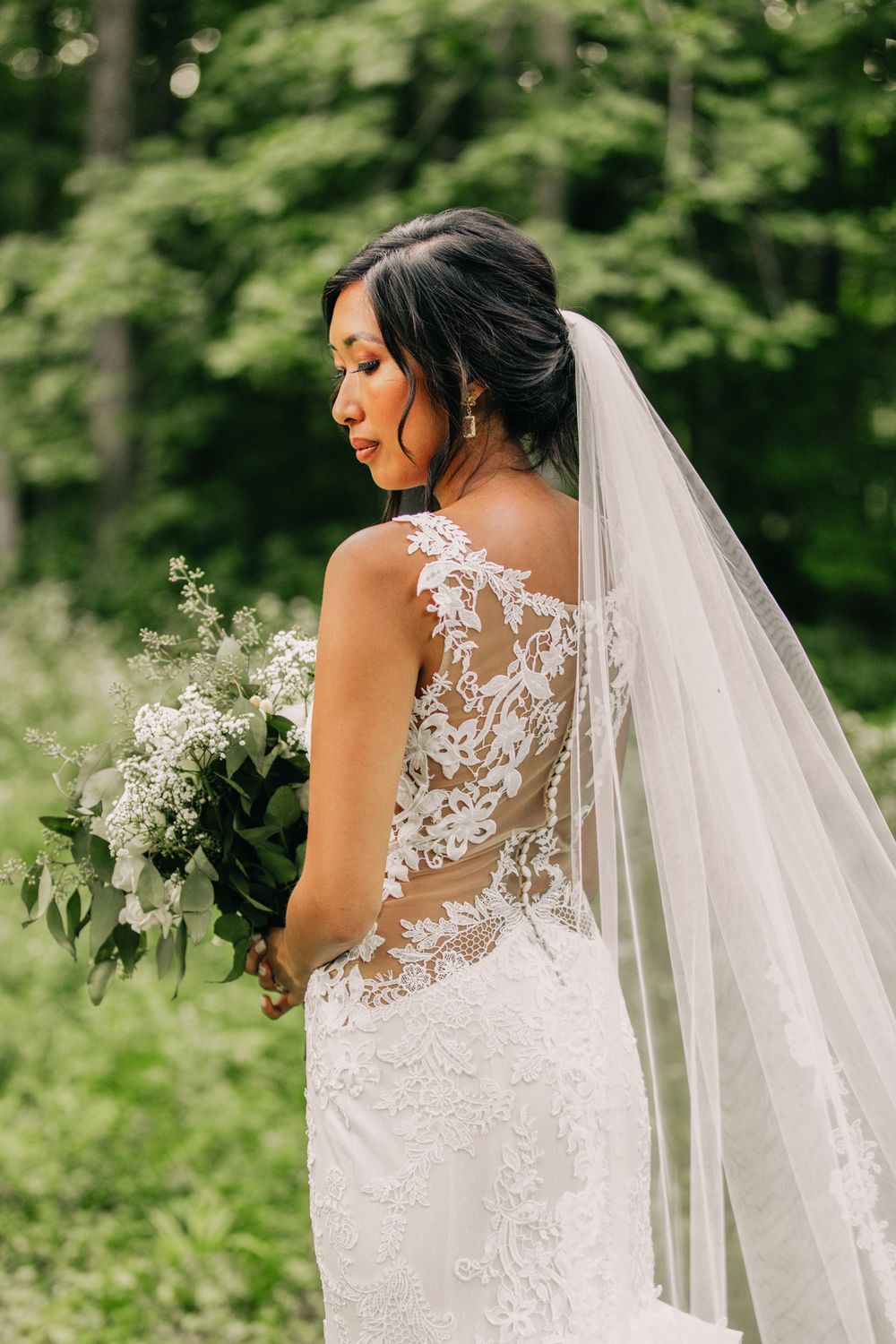 Back view of bride in lace detailed dress with sheer back and flowing veil against forest backdrop.