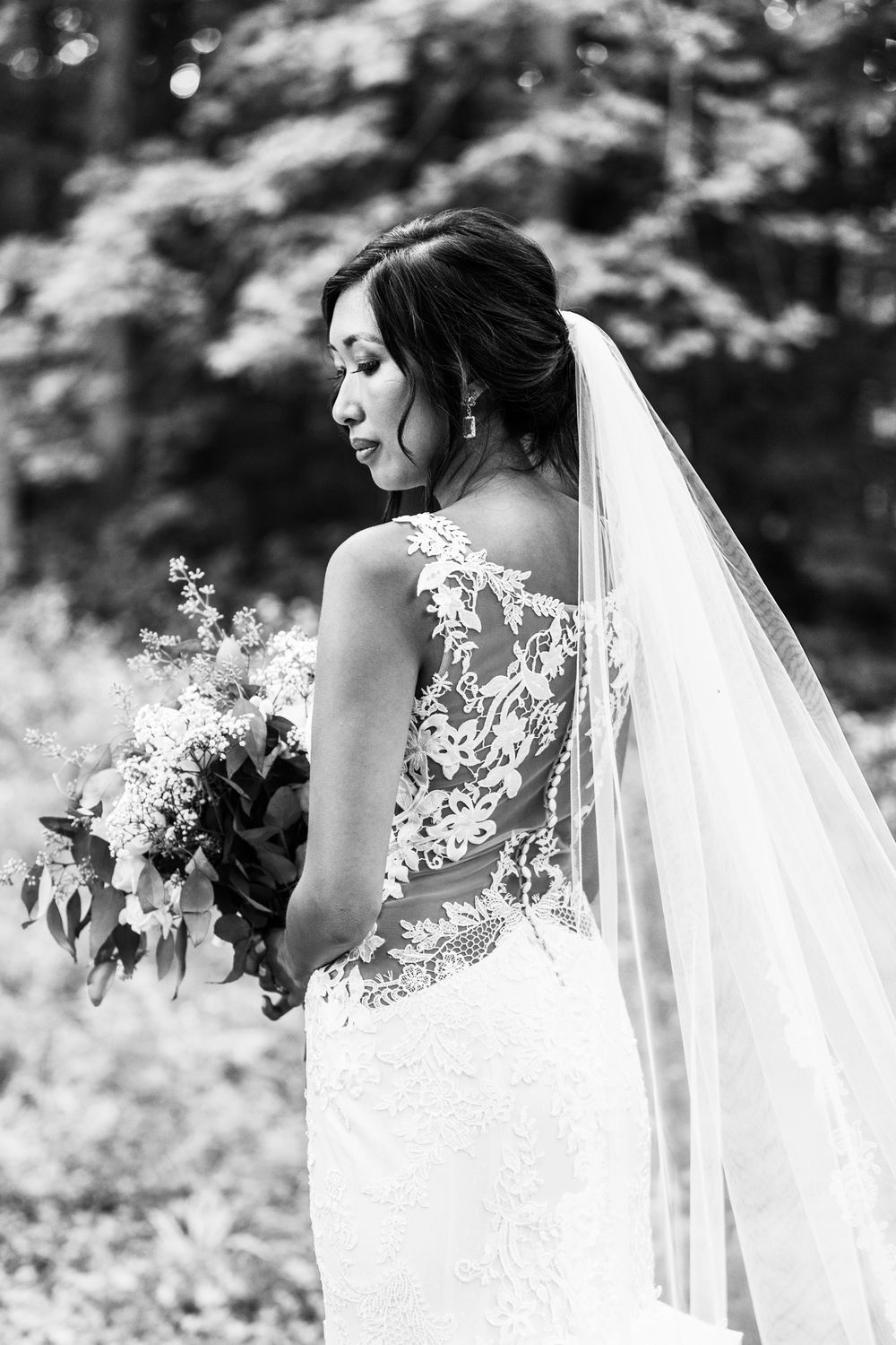 Black and white portrait of bride in lace gown holding bouquet against forest background.