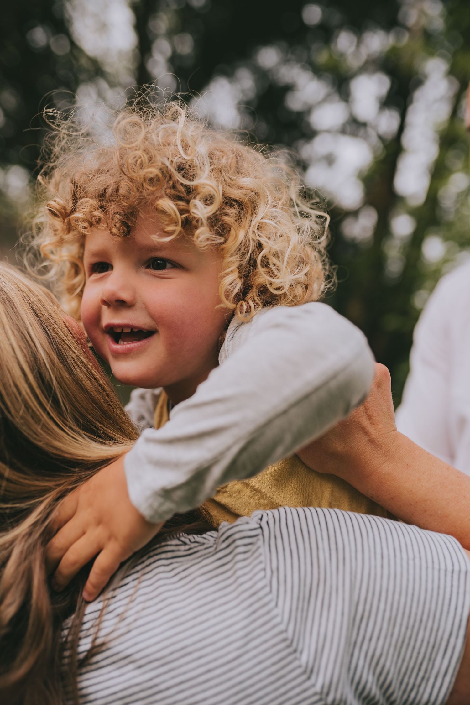 Close-up of joyful toddler with curly hair being held up against sunny outdoor backdrop.