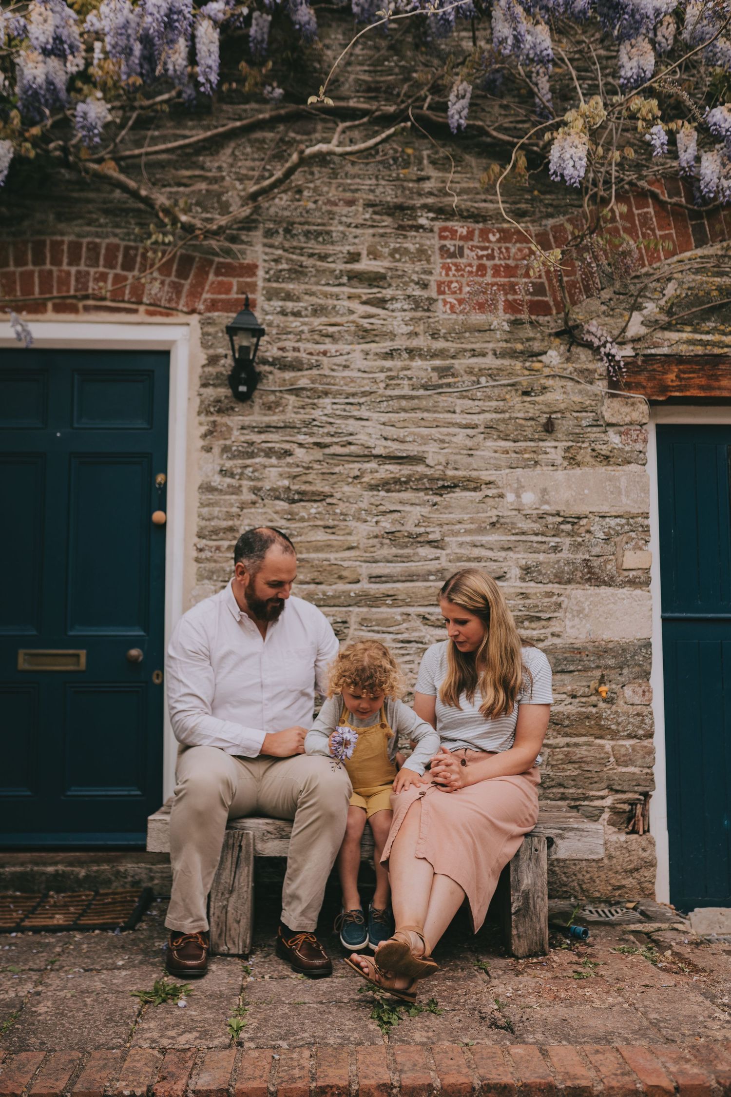 Three people sitting on a stone bench between two green doors with wisteria overhead.