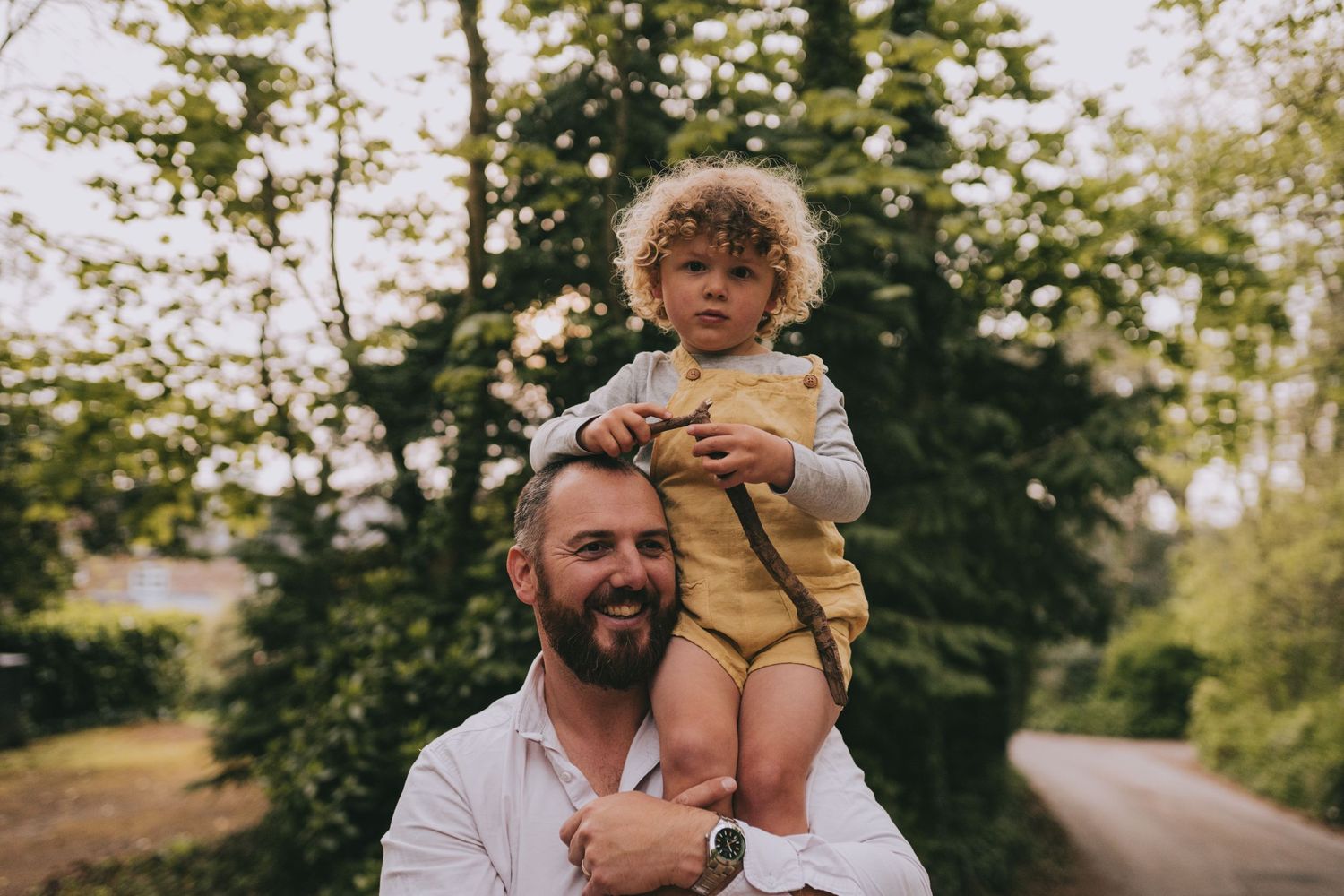 Adult carrying toddler with curly blonde hair on shoulders along wooded path in summer setting.