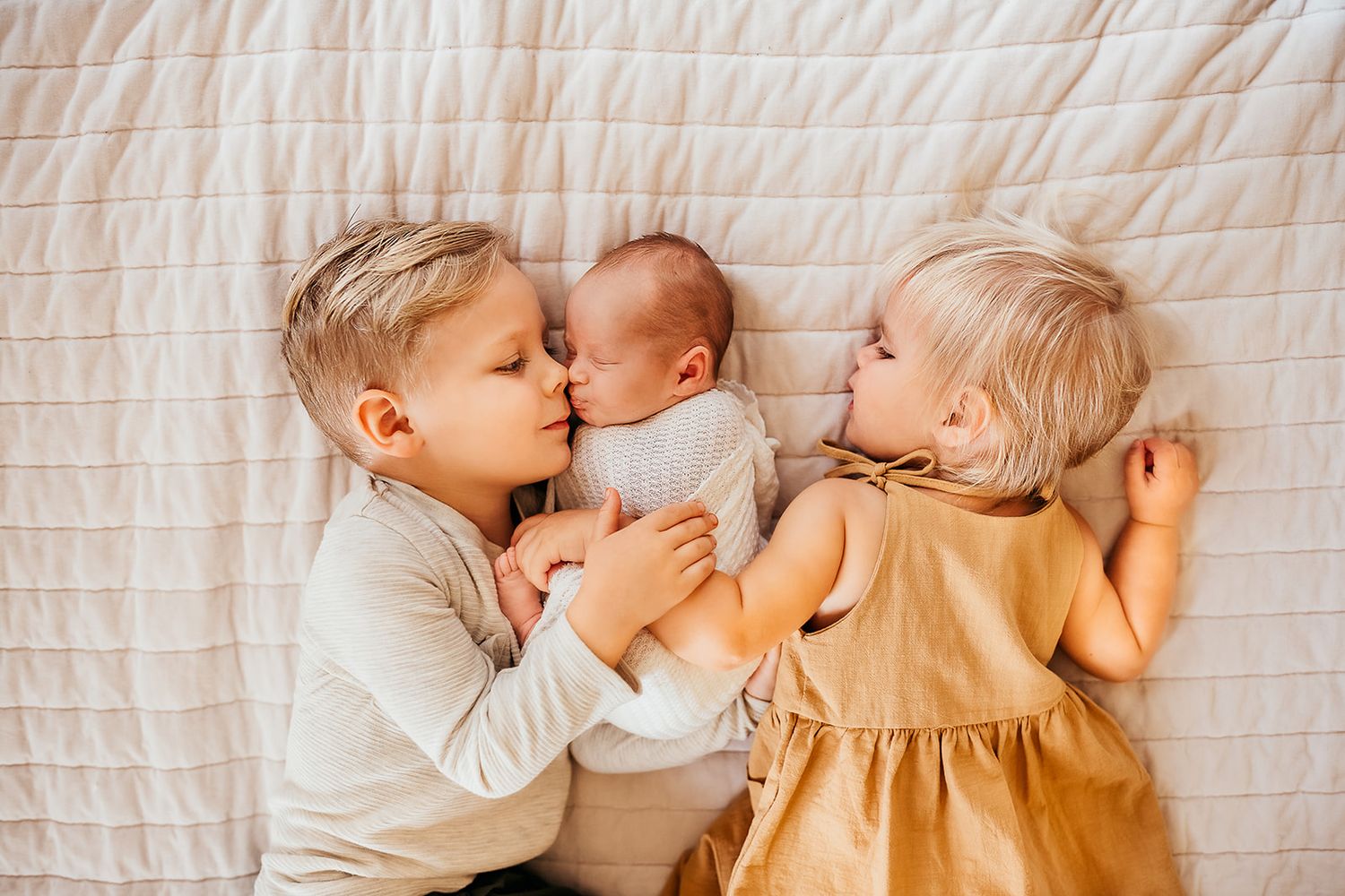 An older brother and sister snuggle their new baby sister on the bed.
