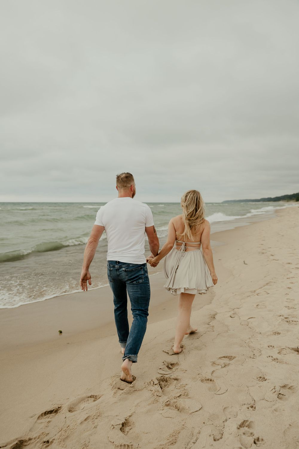 A couple walks hand in hand along the shoreline on an overcast beach day.