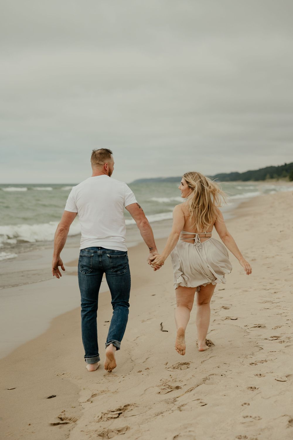 A couple strolls barefoot down a scenic beach with waves and cloudy skies.