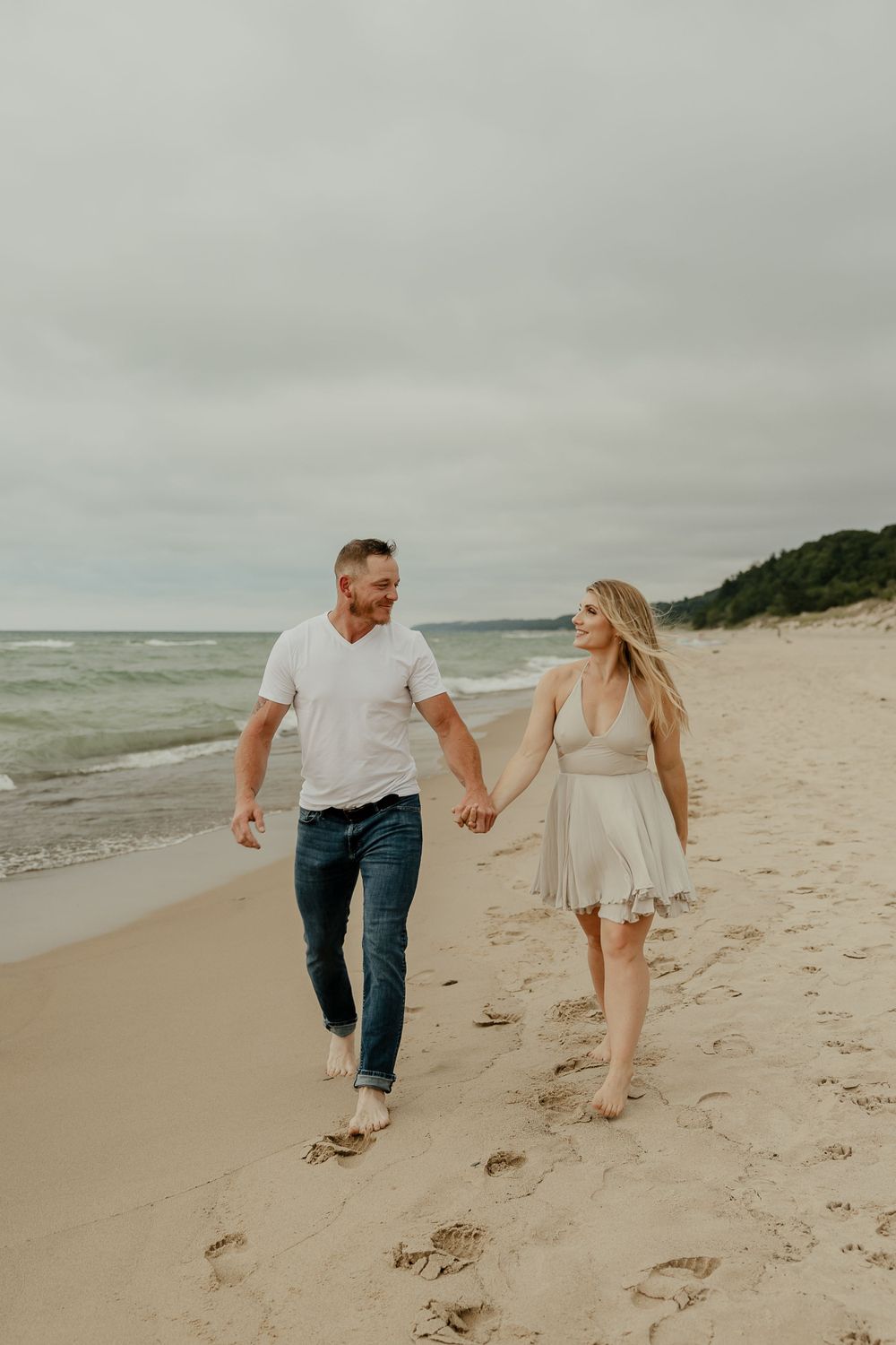 A couple holds hands while walking along a scenic beach with overcast skies.