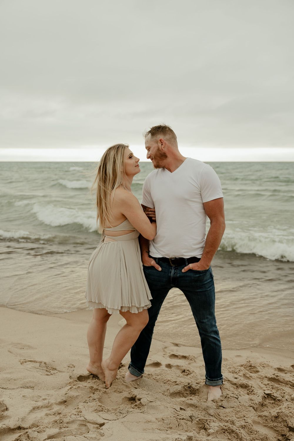 A loving couple stands close together on a sandy beach with waves in the background.
