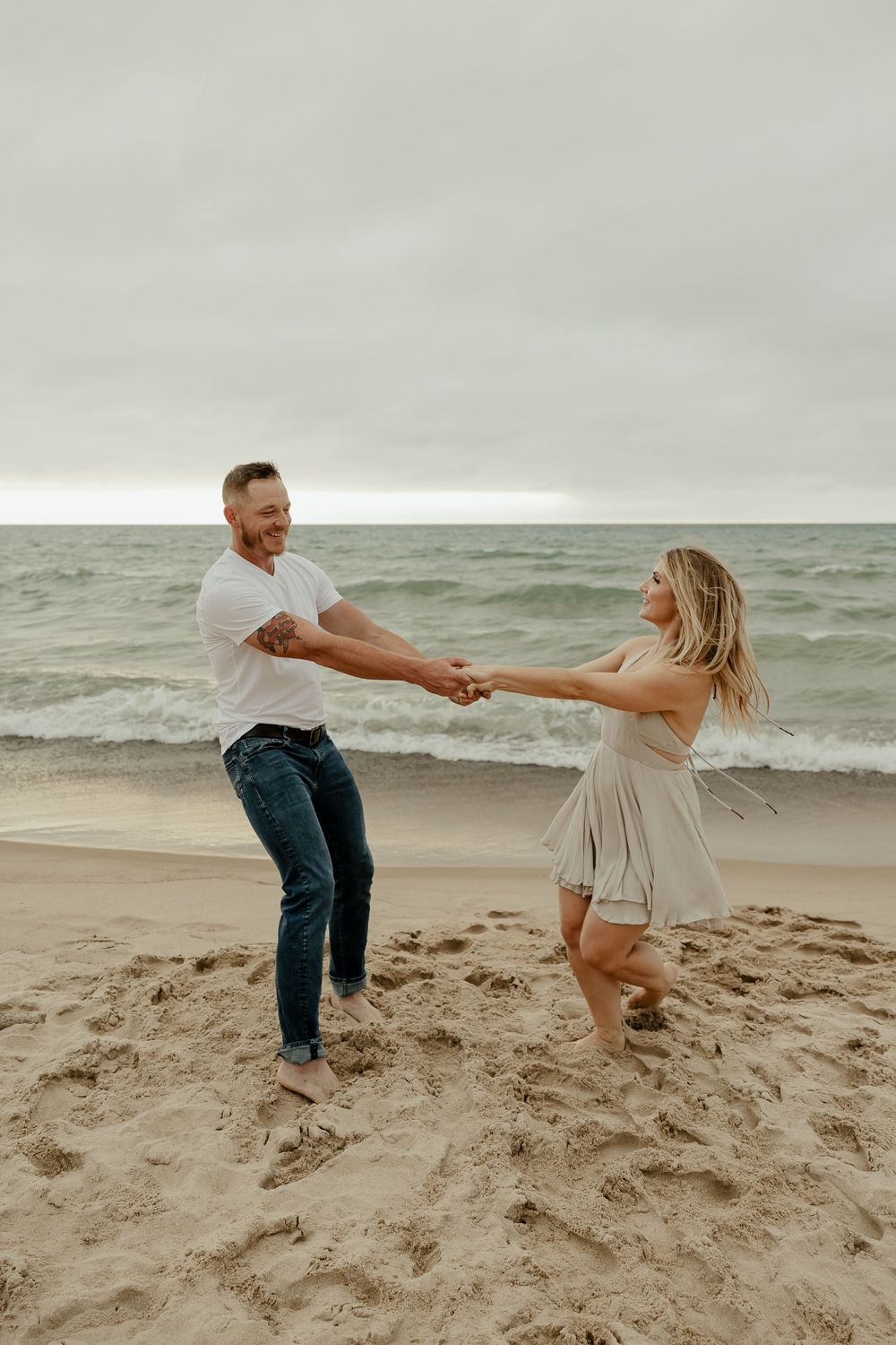 A couple dances playfully on the beach with waves crashing behind them on an overcast day.