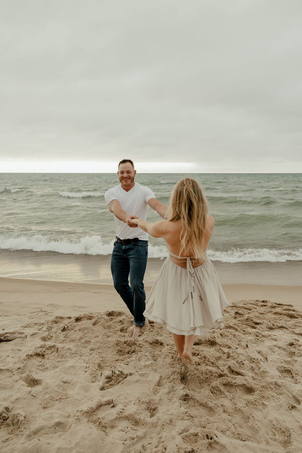 A couple dances and laughs together on a beach with stormy waves in the background.