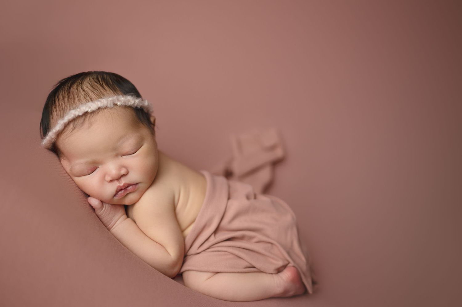 Sleeping newborn in pink wrap against mauve backdrop during professional photo session.