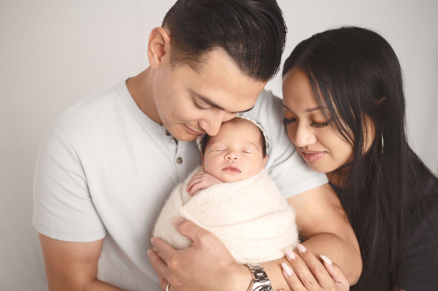 Parents snuggling with their sleeping newborn baby wrapped in white.